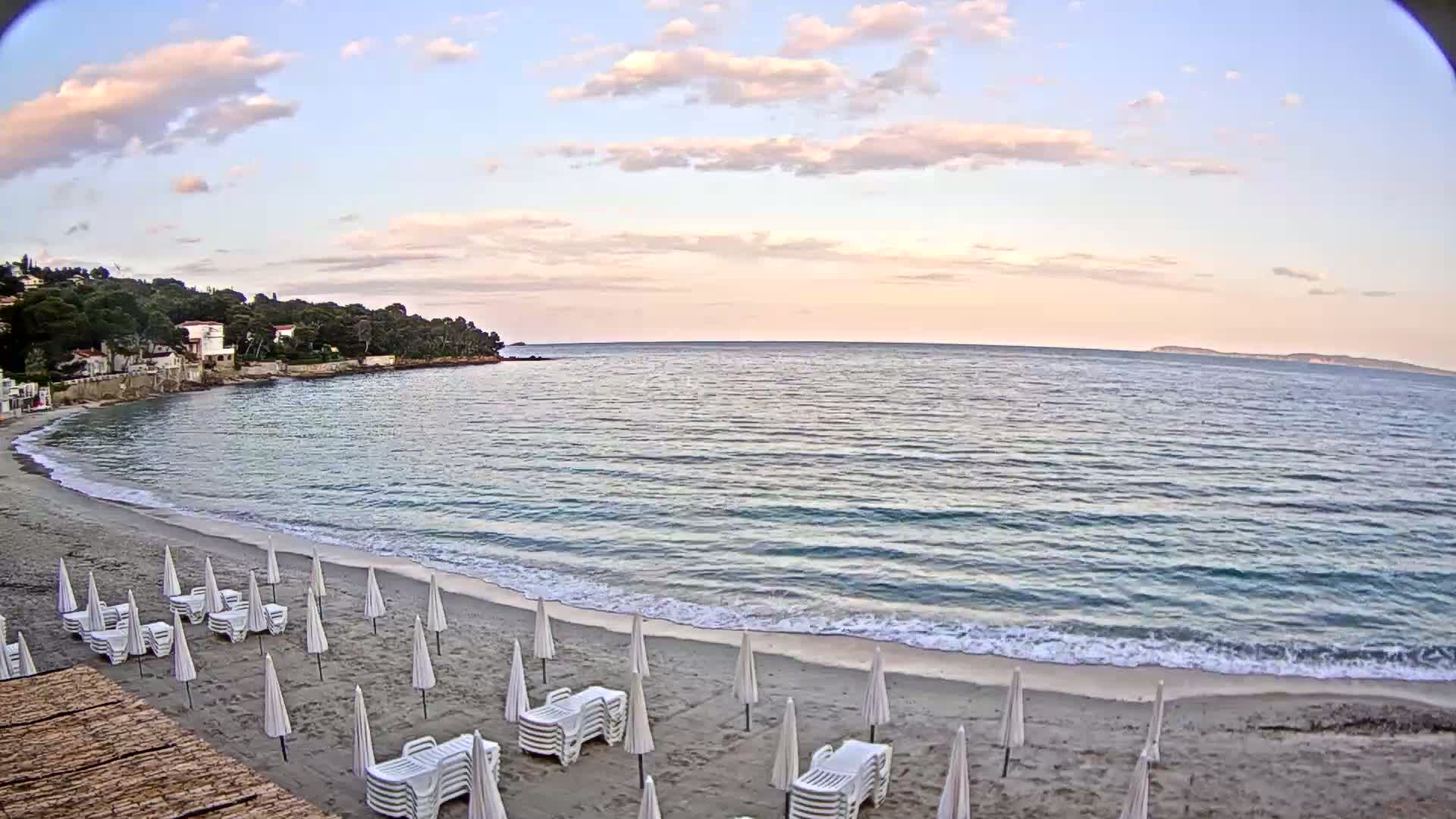 A sandy beach with rows of white lounge chairs and umbrellas under a partly cloudy sky overlooks a calm sea with gentle waves.