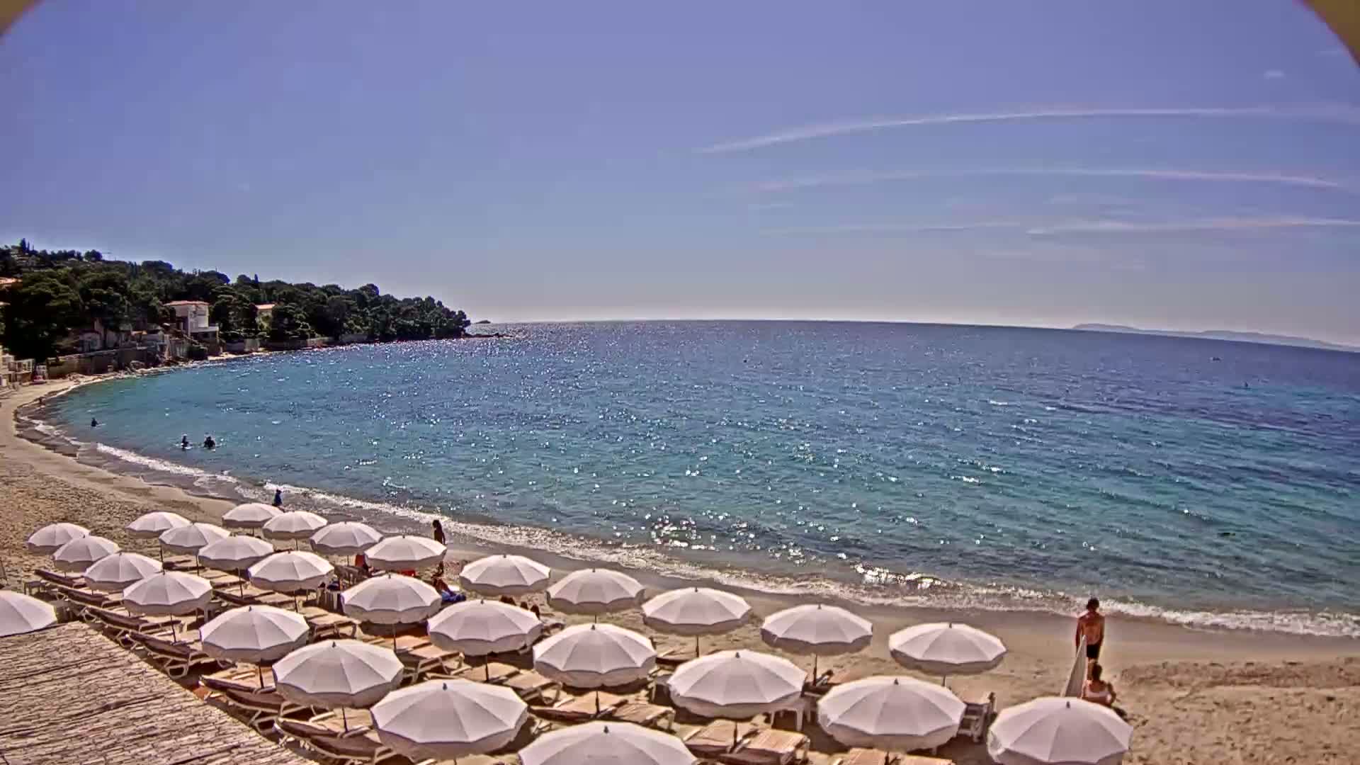 A sandy beach with white umbrellas and lounge chairs lines a calm, clear turquoise sea under a sunny sky.