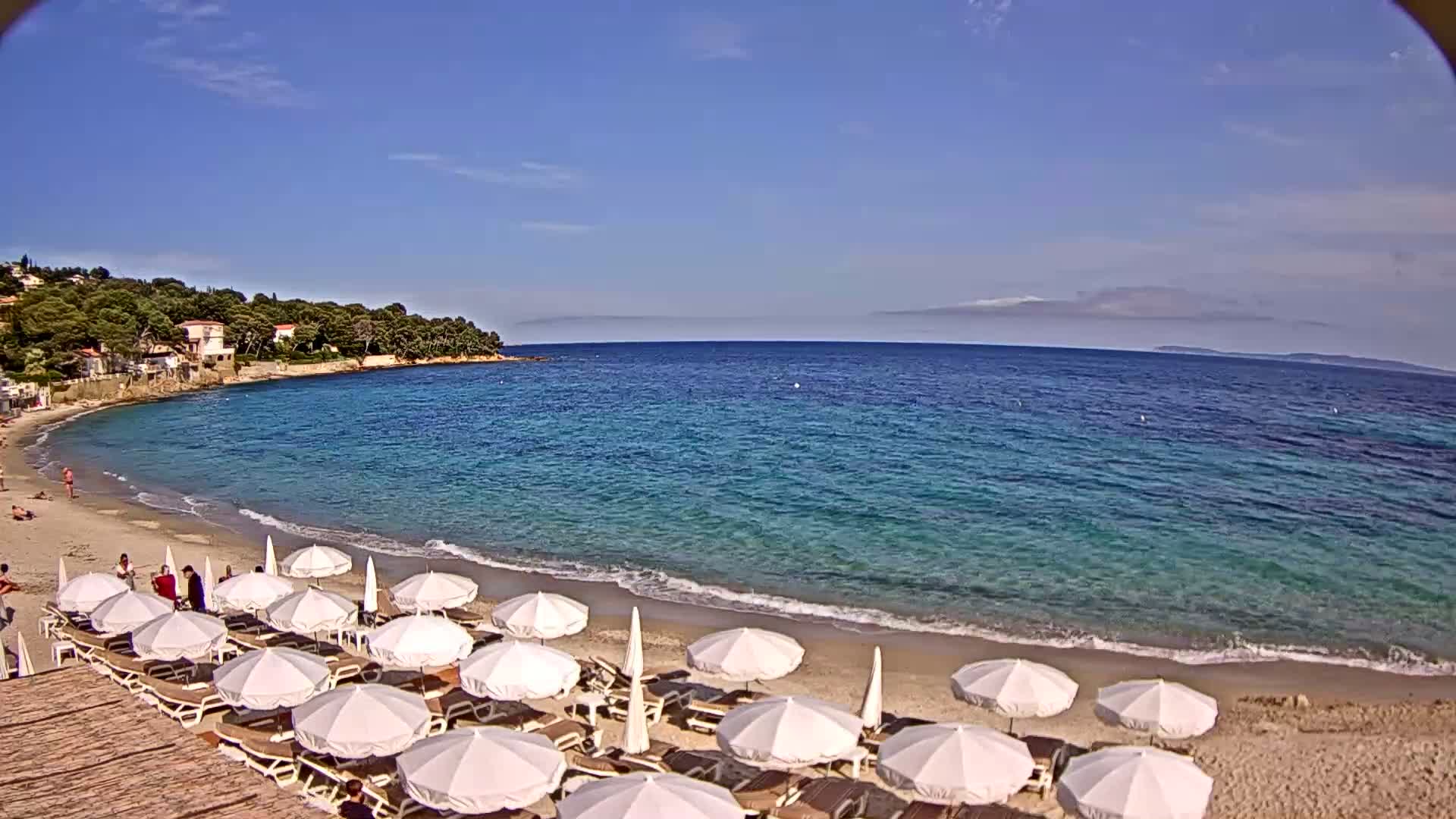 A sandy beach with white umbrellas and lounge chairs lines a calm, clear turquoise sea under a bright, sunny sky.