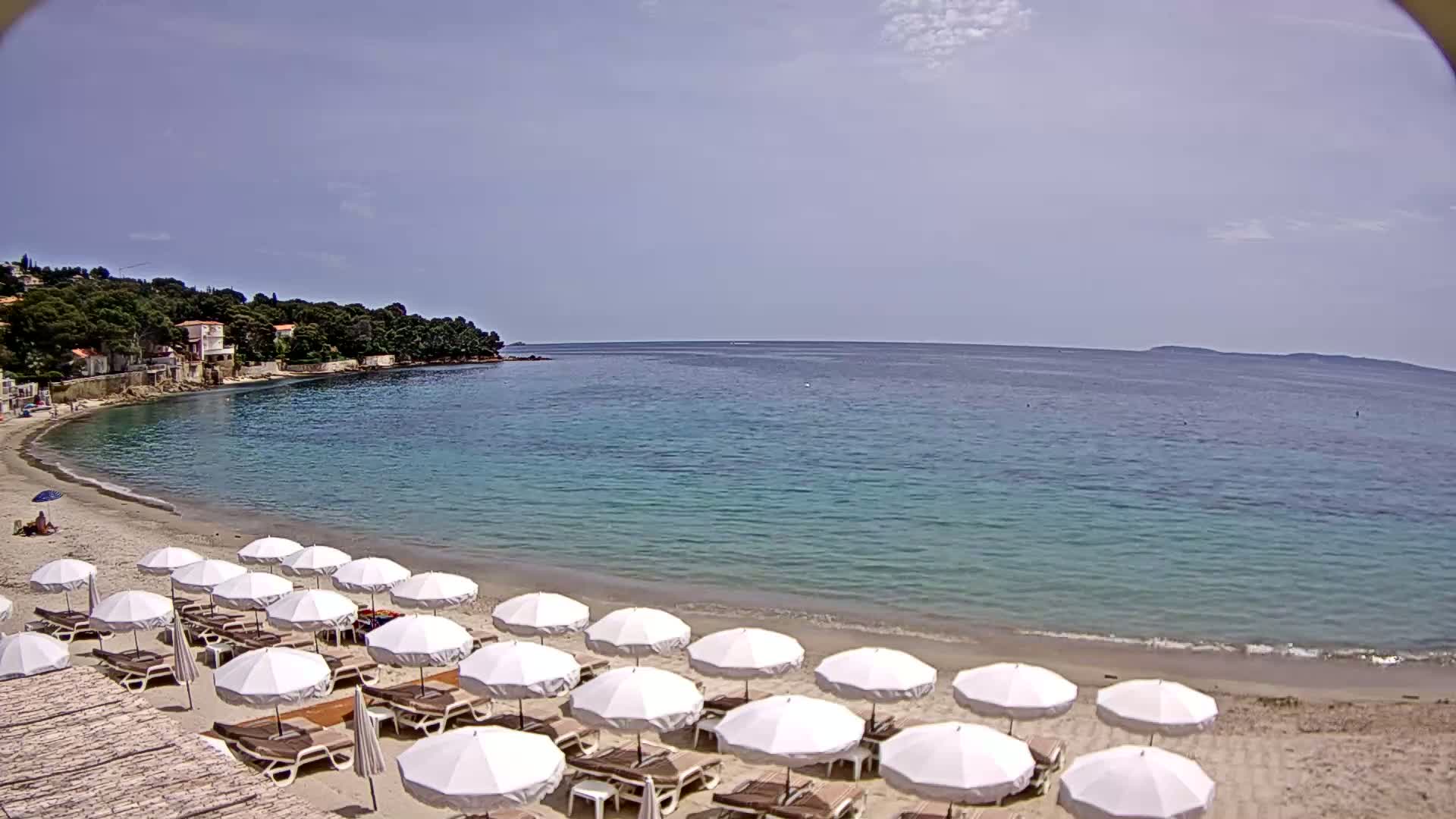 A sandy beach with numerous white umbrellas and lounge chairs lines a calm, turquoise bay under a mostly clear sky.