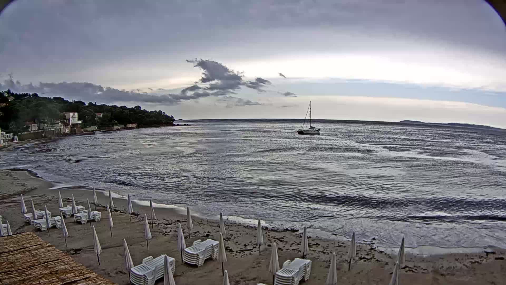 A mostly cloudy day shows a sailboat on the water near a sandy beach with rows of closed white beach umbrellas and lounge chairs.