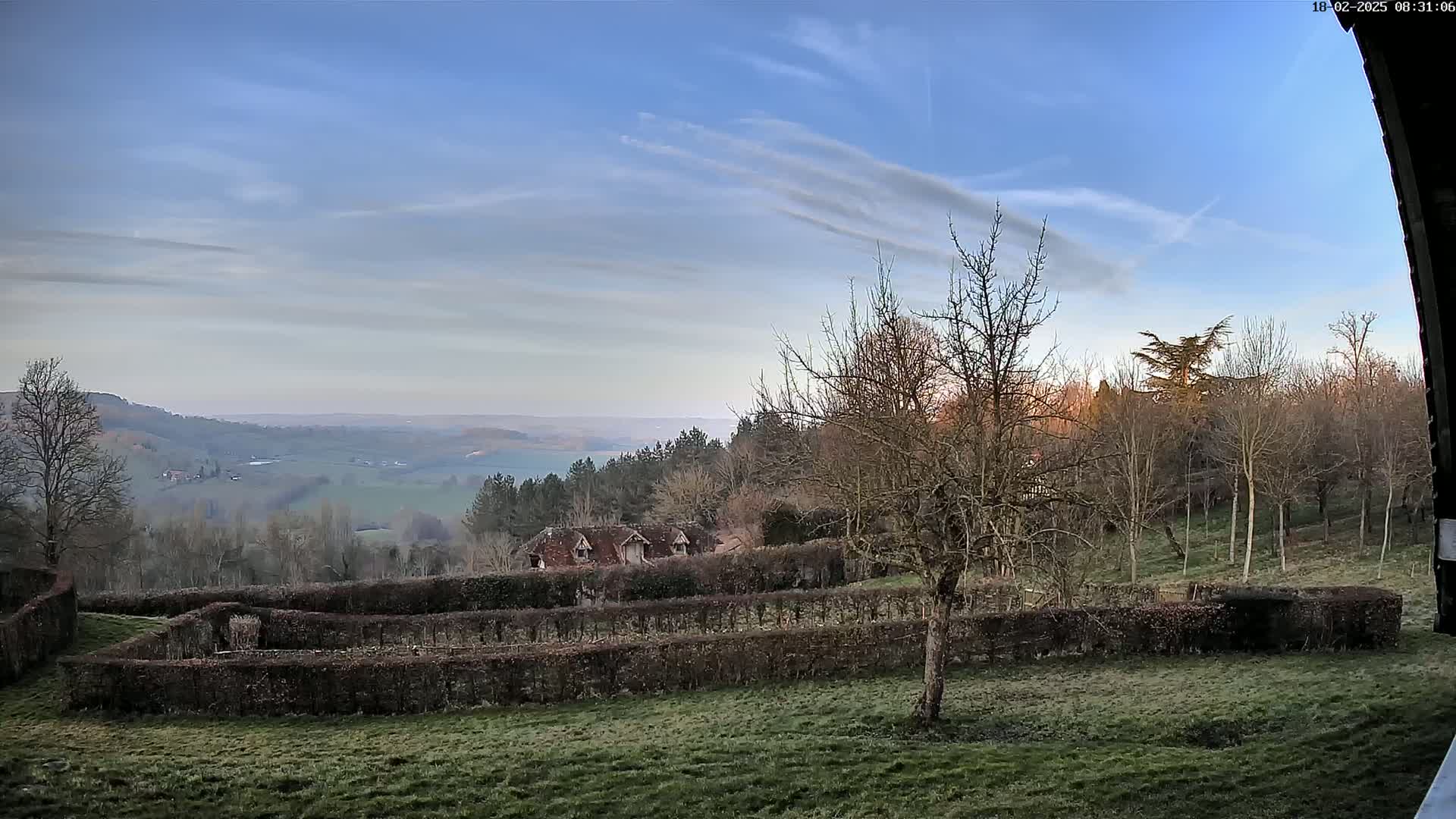 A sprawling, hilly landscape under a mostly clear, pale blue sky is viewed from a slight elevation, showing leafless trees, a building partially obscured by hedges, and a large, meticulously trimmed hedge maze in the foreground.