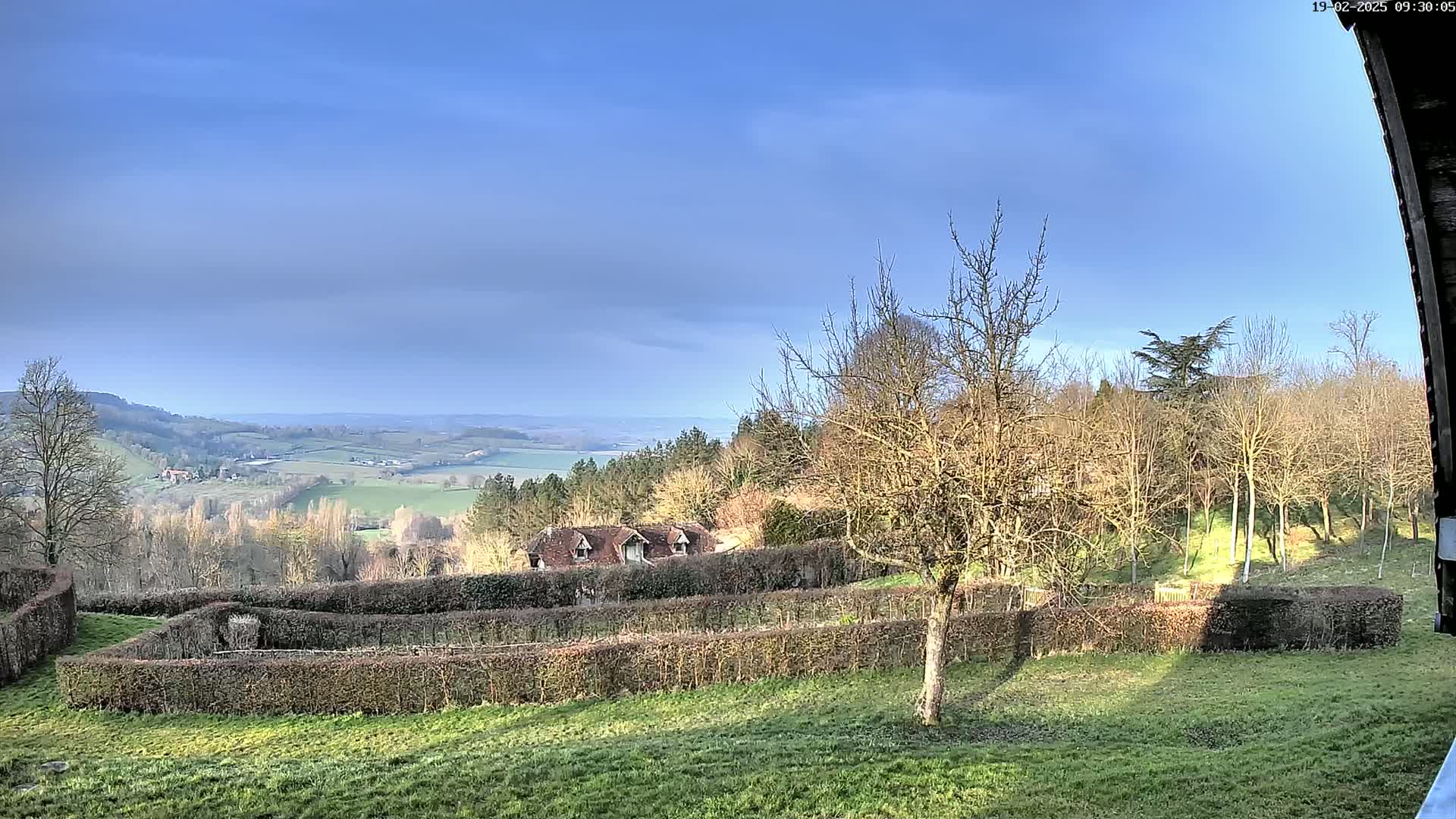 A sunny, partly cloudy day reveals a rolling green landscape with a building nestled among leafless trees and meticulously trimmed hedges, all viewed from a slight elevation.