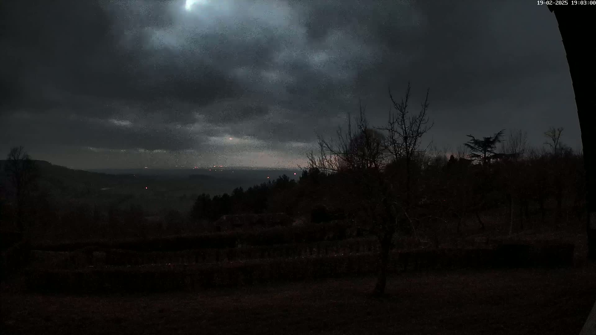 A dark, overcast night scene shows a hilly landscape with silhouetted trees and distant city lights visible on the horizon.