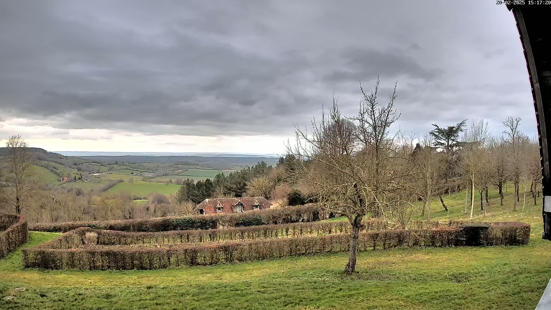 A rural landscape under a cloudy sky shows a building nestled among hedgerows and trees overlooking a vast, rolling green valley.