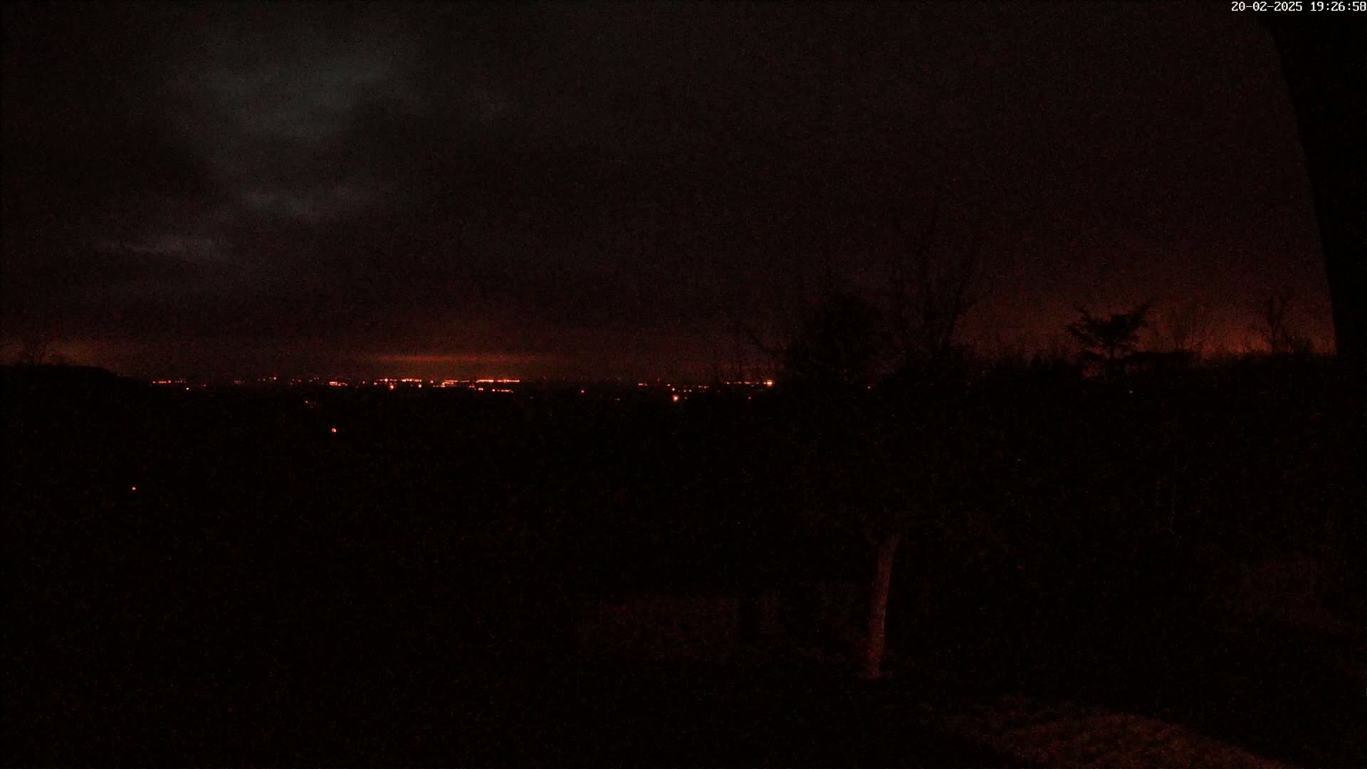 A nighttime view of a distant city skyline illuminated with orange lights under a dark, cloudy sky.