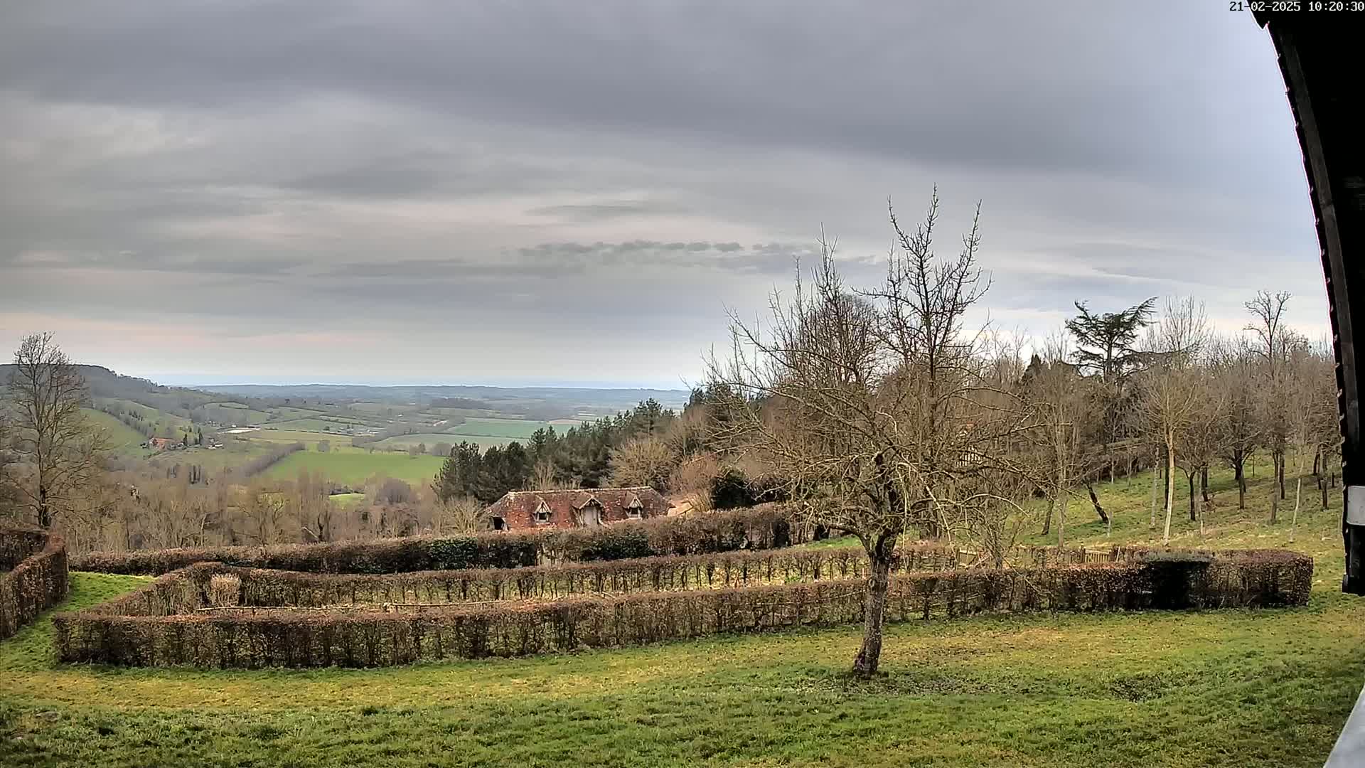 A sprawling, verdant valley under a cloudy sky is visible from a vantage point overlooking a hedged garden and a low, rustic building.