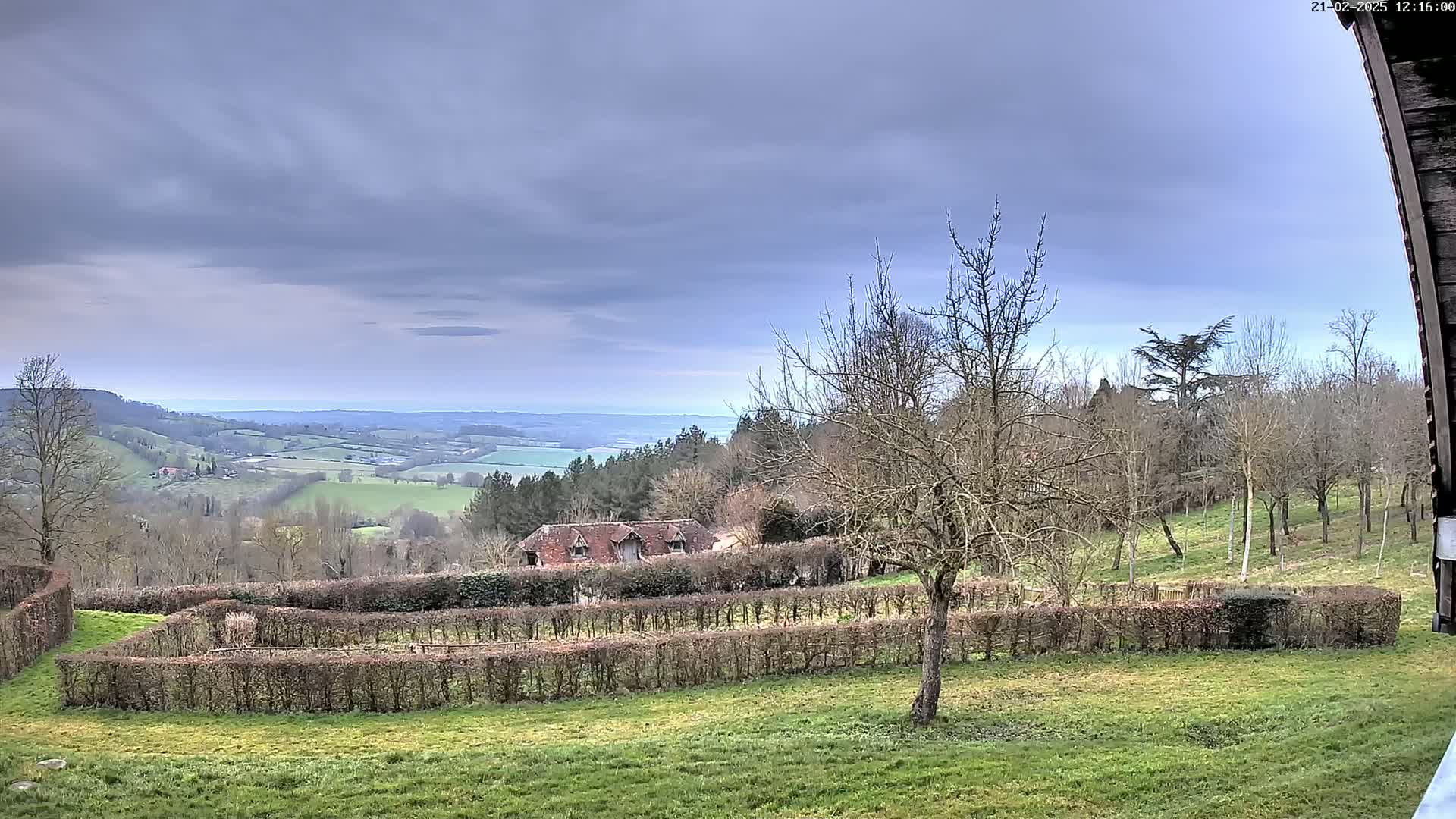 A sprawling, hilly landscape under a cloudy sky features a building nestled among bare trees and neatly trimmed hedges, overlooking a distant body of water.