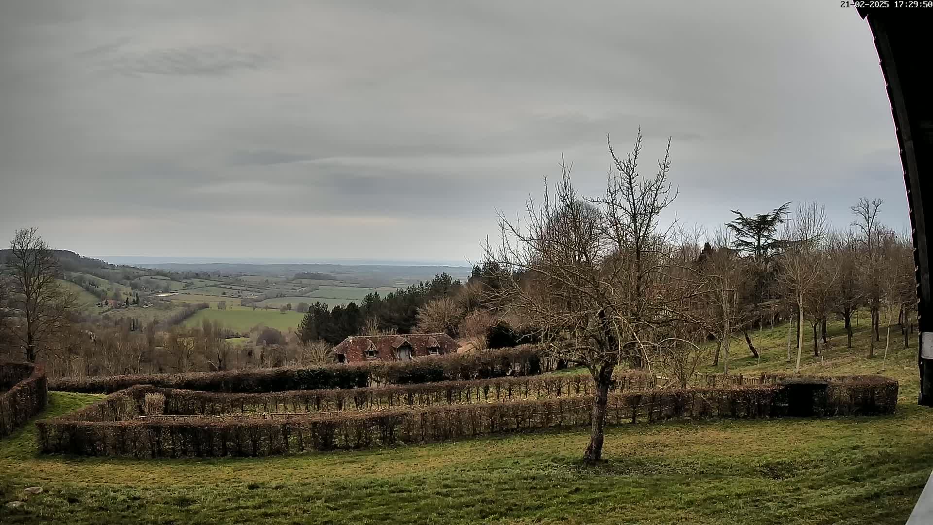 A sprawling landscape under a gray sky shows a stone building nestled amongst leafless trees and neatly trimmed hedges, overlooking a vast valley.