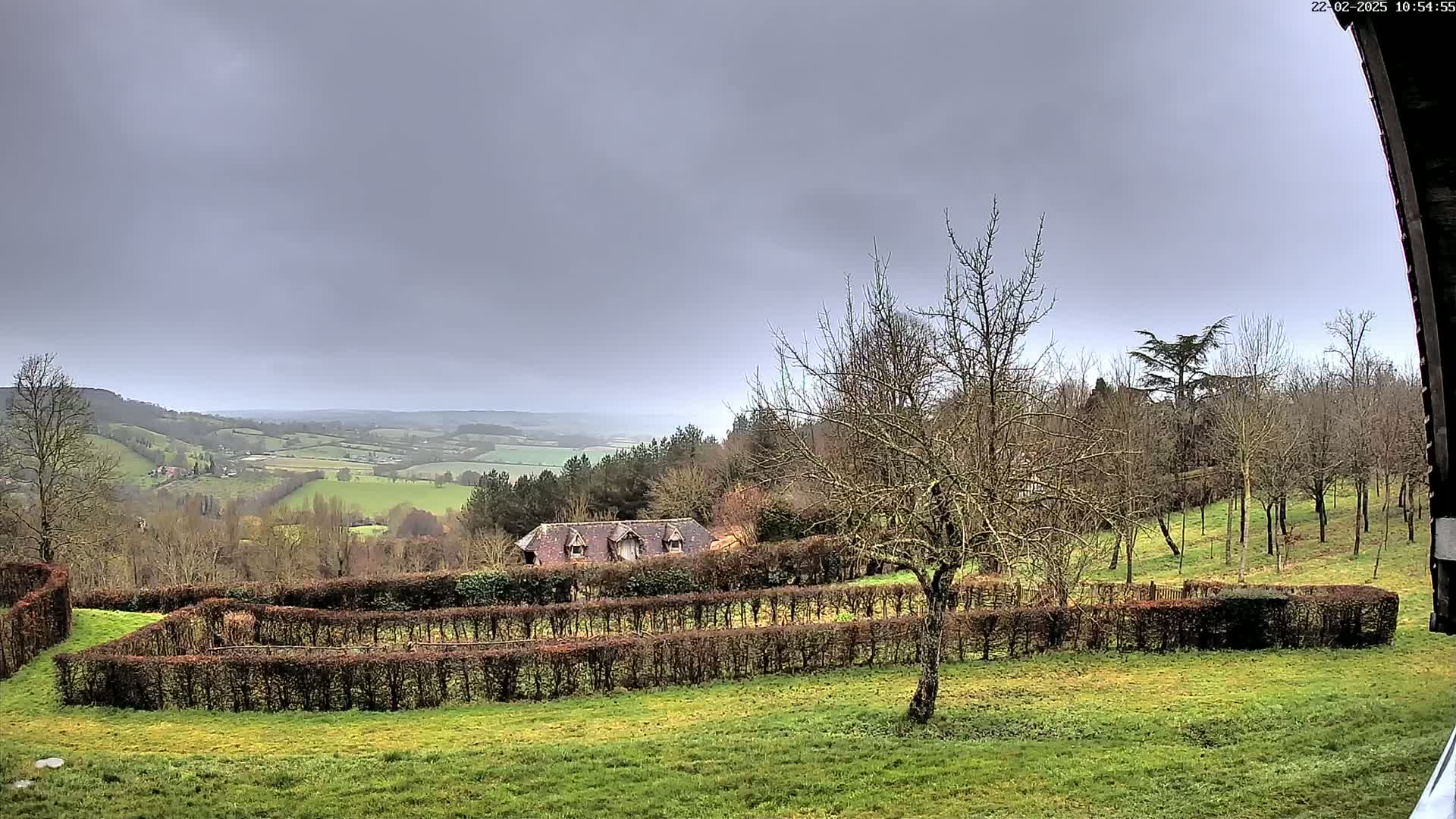 A sprawling, overcast landscape features a house nestled behind a series of hedges, overlooking a valley dotted with trees and fields.