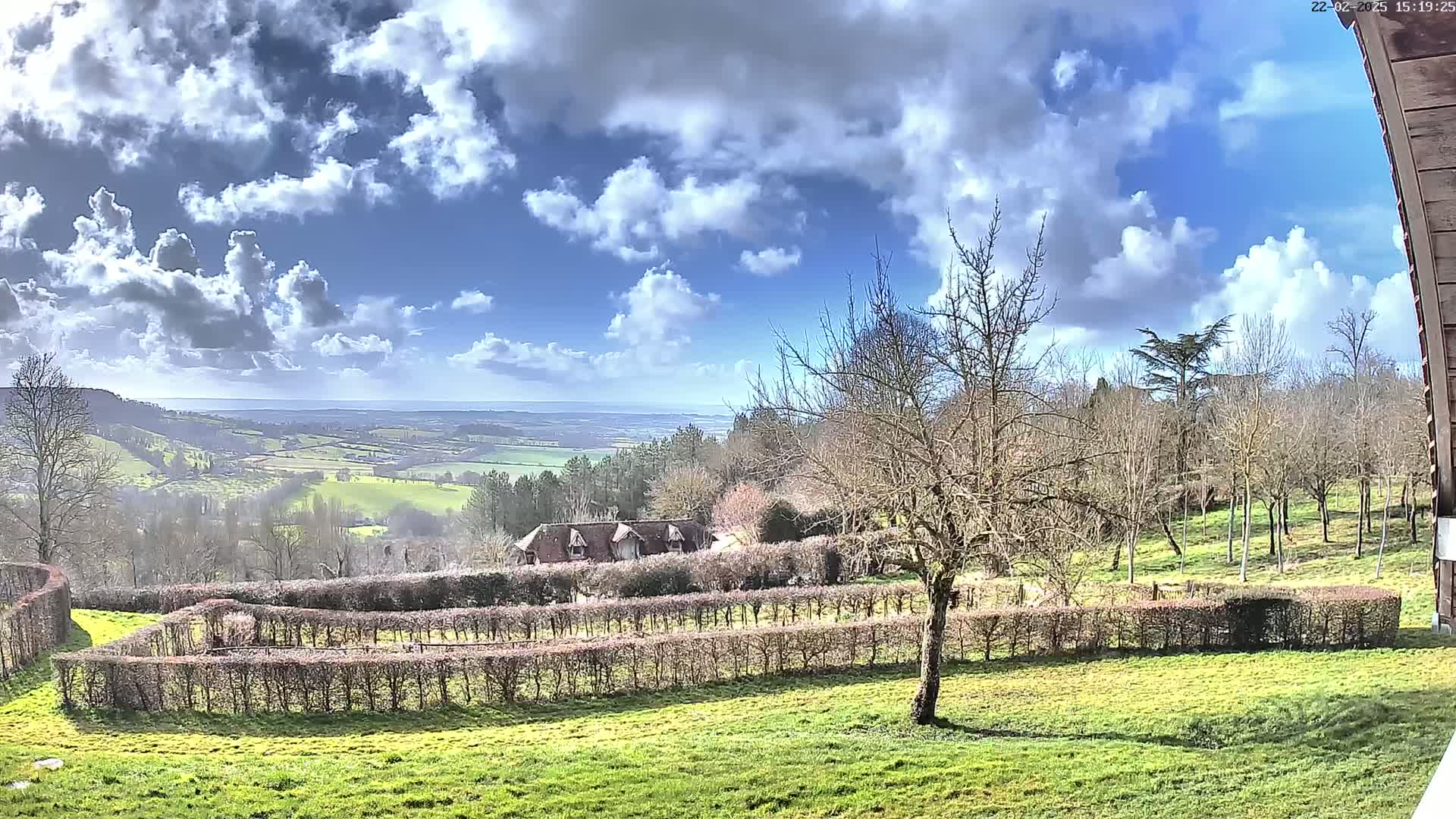A partly cloudy, sunny day overlooks a sprawling green landscape with neatly trimmed hedges, a distant house, and a view of the sea in the background.