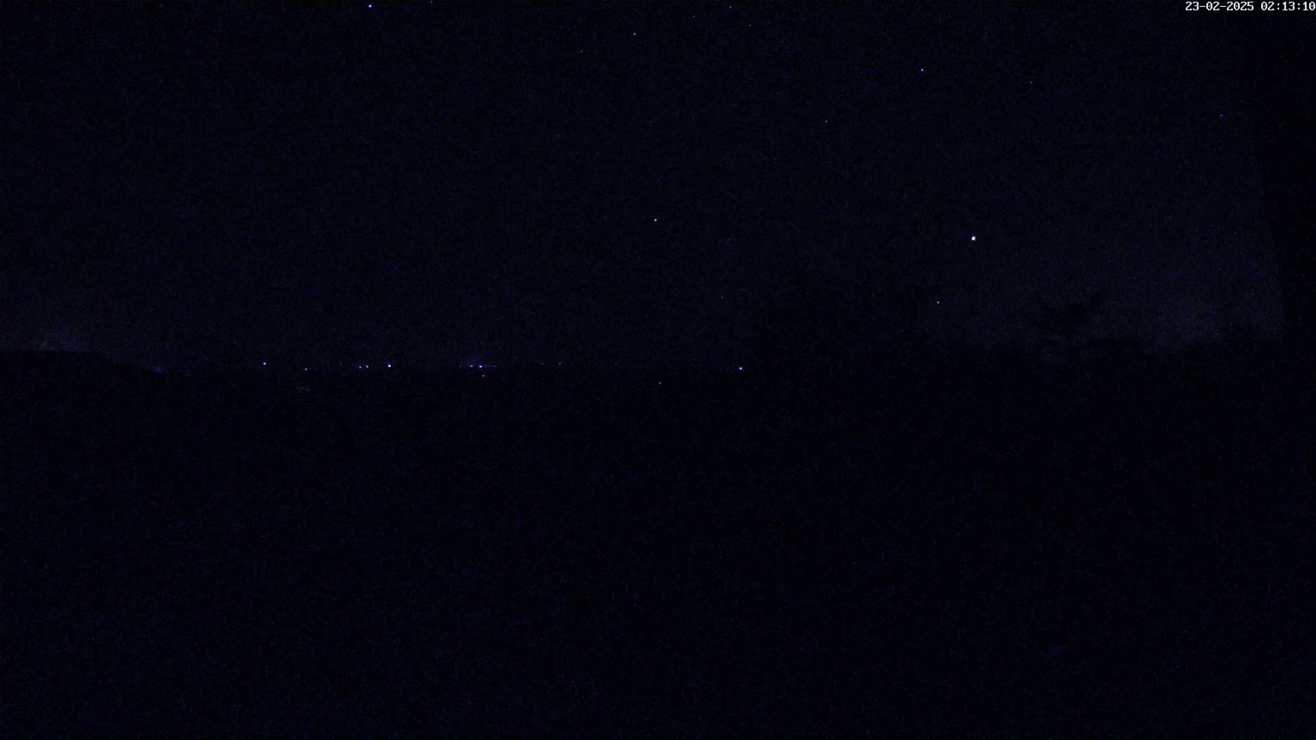 A nighttime view of a dark landscape with a few distant lights and a clear, starry sky.