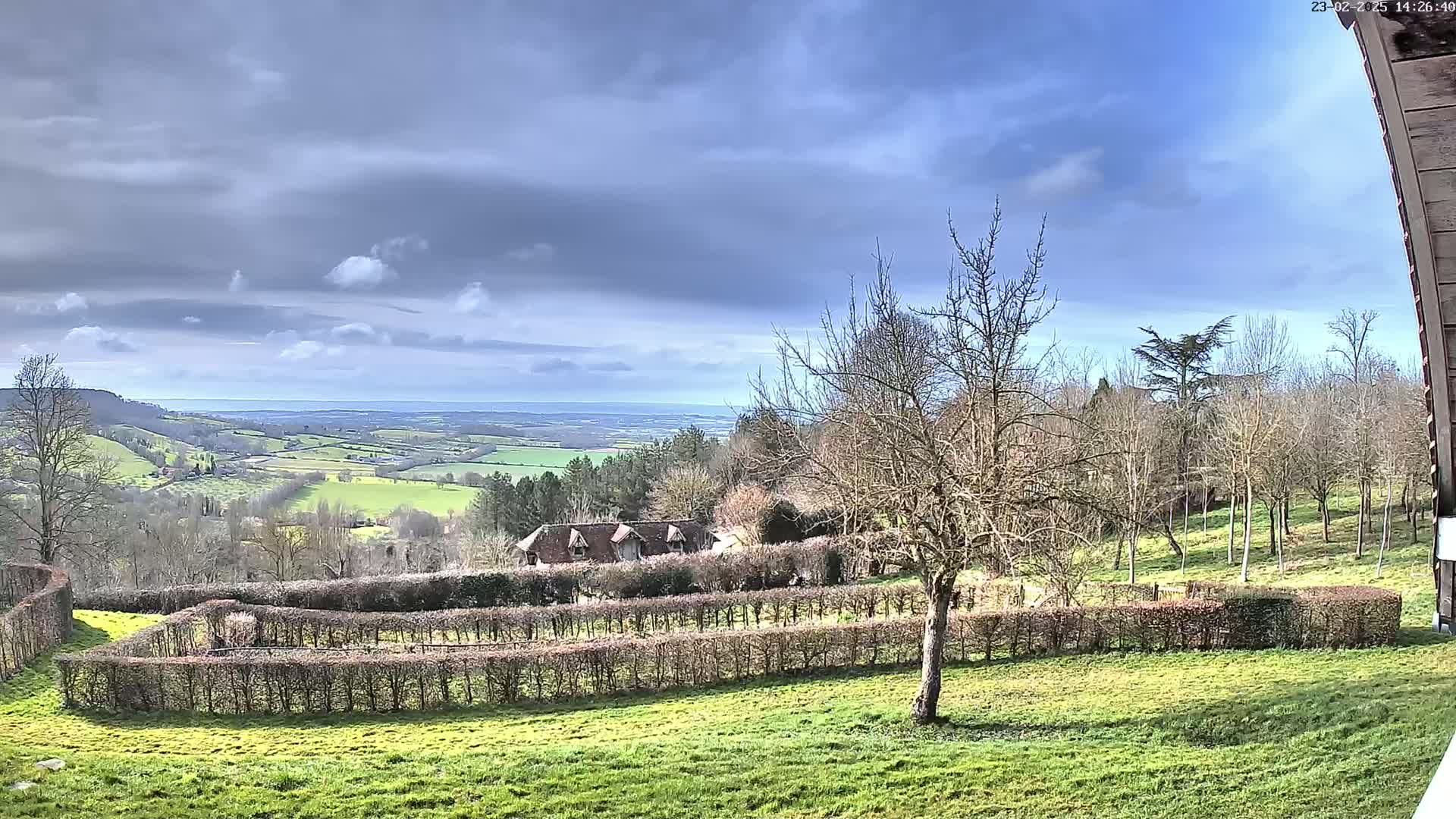 A partly cloudy sky overlooks a sprawling green valley with a house nestled behind a winding hedge, surrounded by bare winter trees.