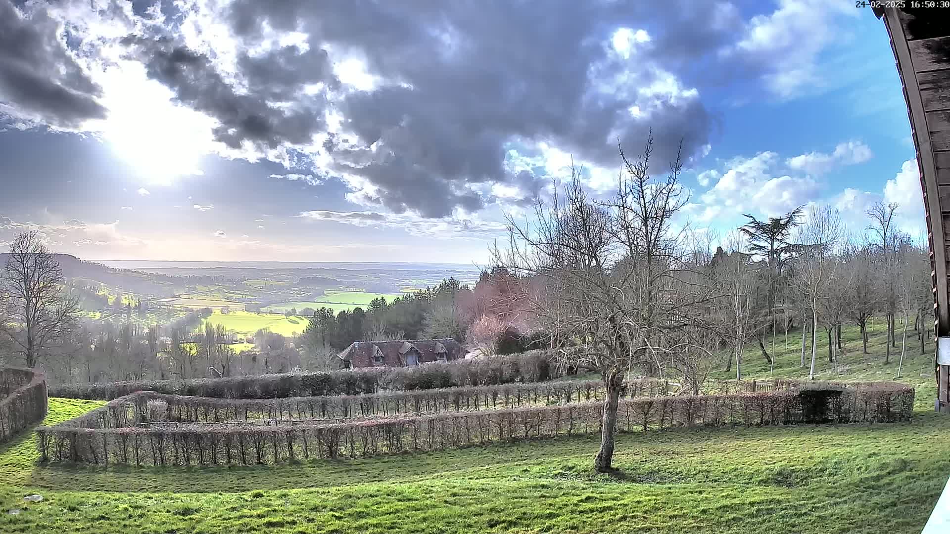A partly sunny day reveals a sprawling green valley with a distant body of water, overlooked by a house nestled amongst leafless trees and neatly trimmed hedges.