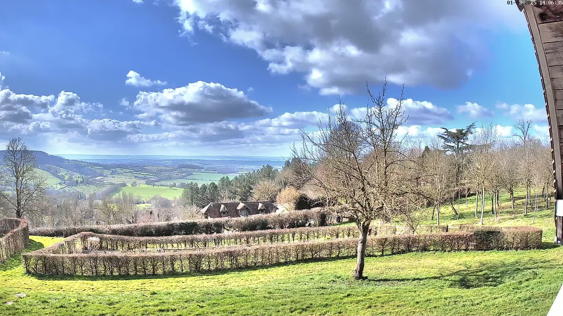A partly cloudy day reveals a panoramic view of a rolling green landscape dotted with trees and a building nestled behind a meticulously trimmed hedge maze.