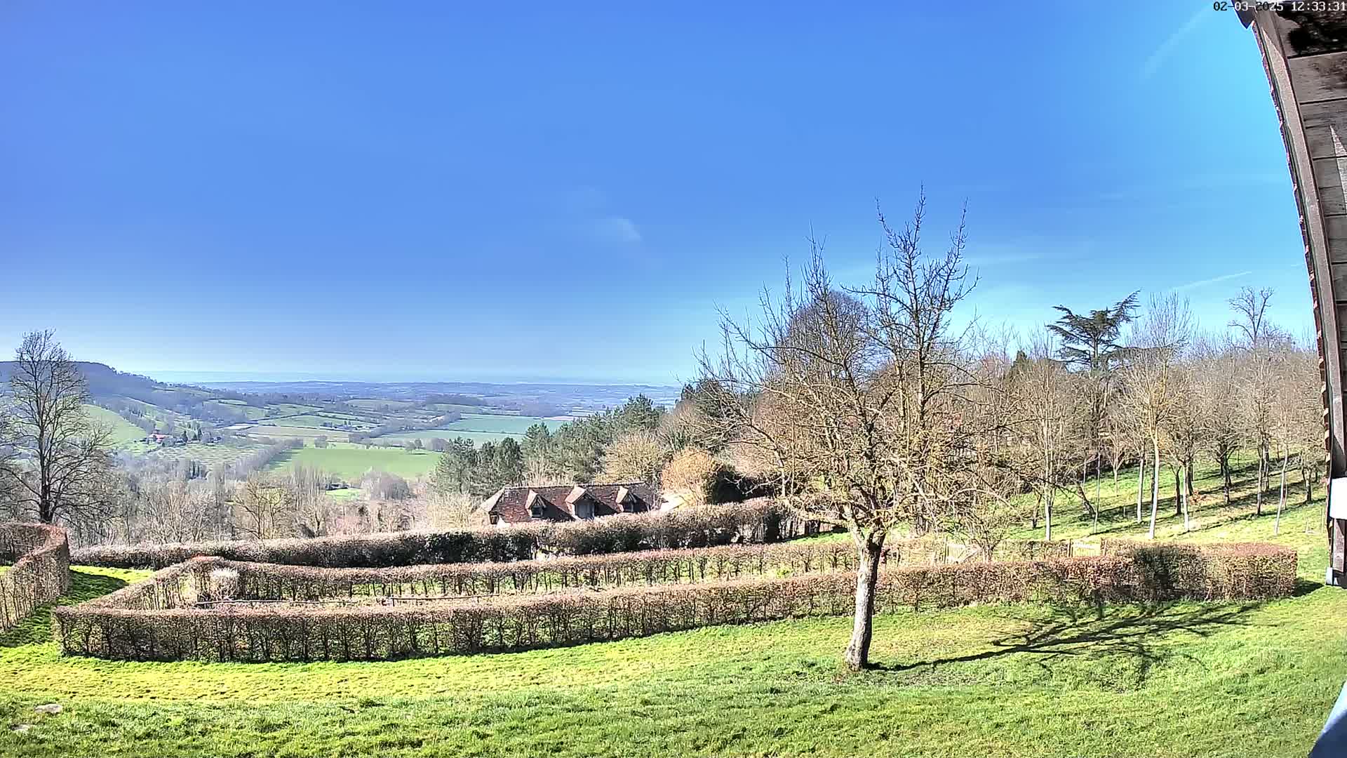 A sunny day reveals a sprawling green landscape, featuring a long hedgerow, a distant house, and a clear view of a valley extending to a horizon where the sea is visible.