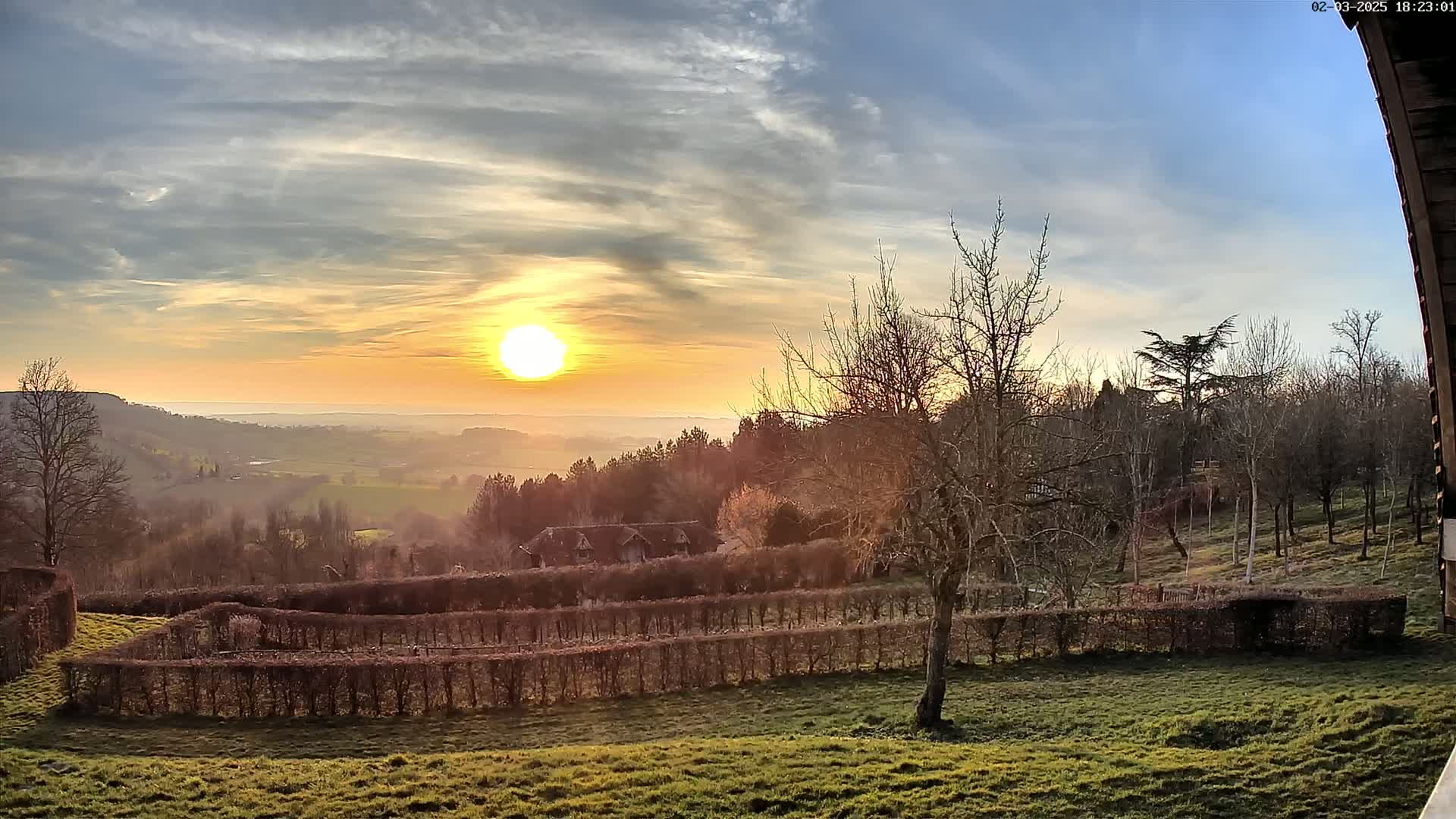 A sunset over a vast, hazy valley with a line of trimmed hedges in the foreground and leafless trees dotting the landscape.