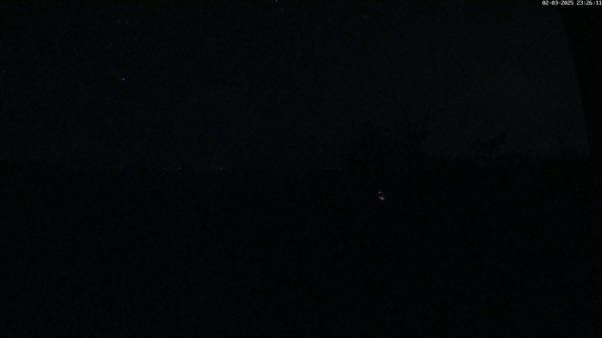 A nighttime, dark scene shows a few faint lights in the distance under a mostly clear, starlit sky.