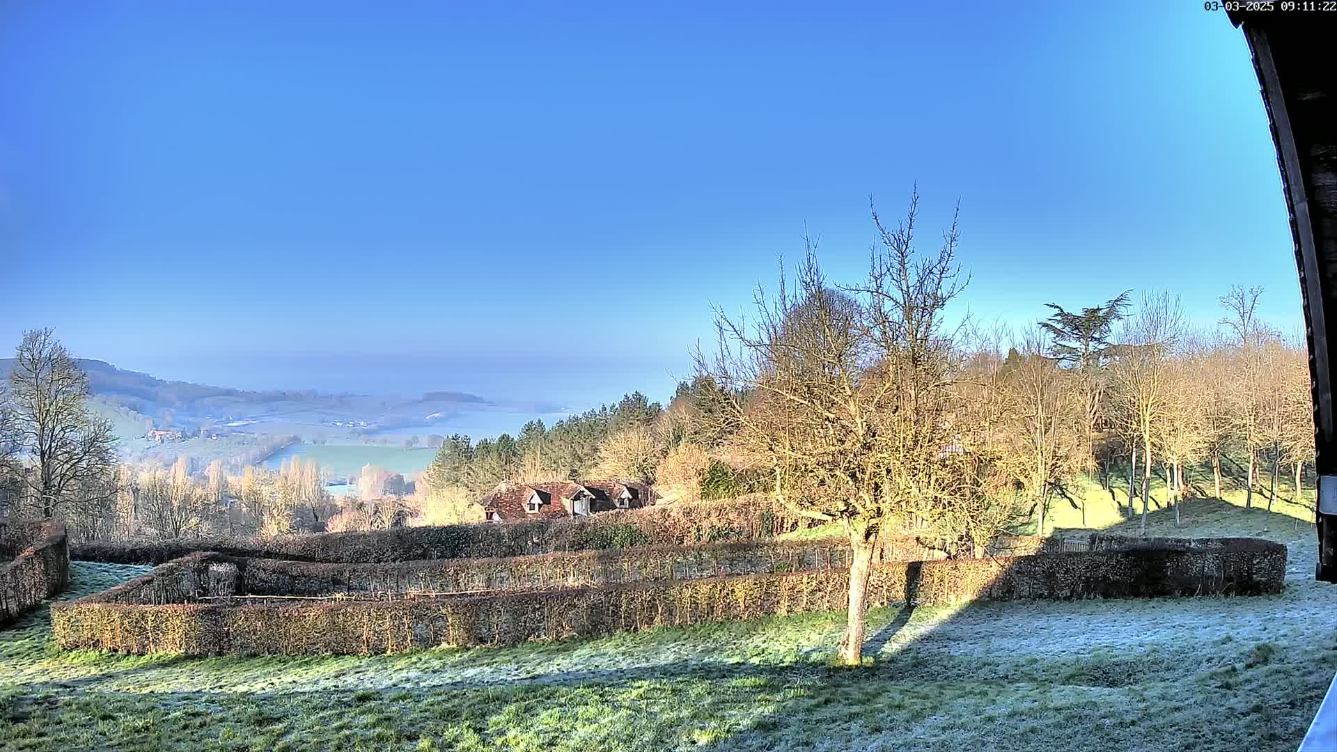A frost-covered field with trimmed hedges and leafless trees overlooks a valley shrouded in morning mist under a clear blue sky.