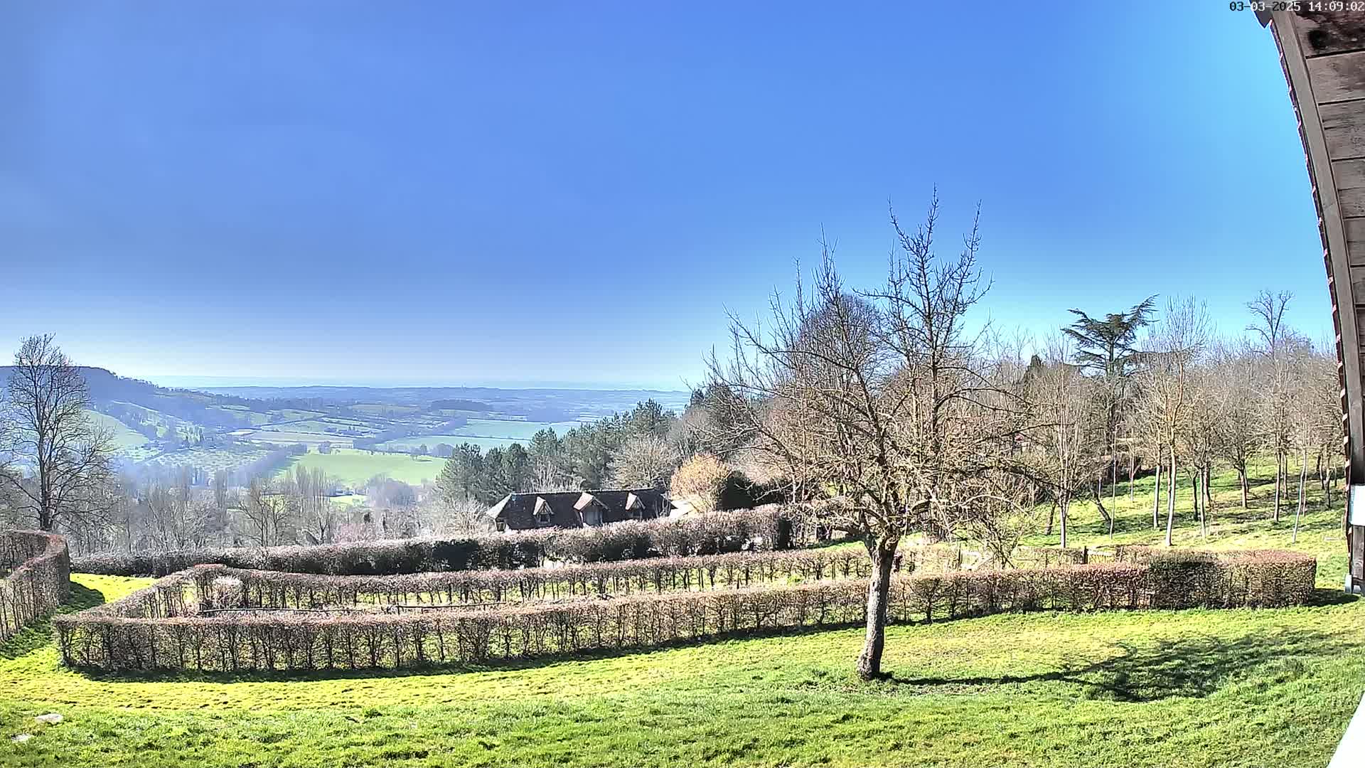 A sprawling green landscape under a clear blue sky features a building nestled among meticulously trimmed hedges, overlooking a distant valley.
