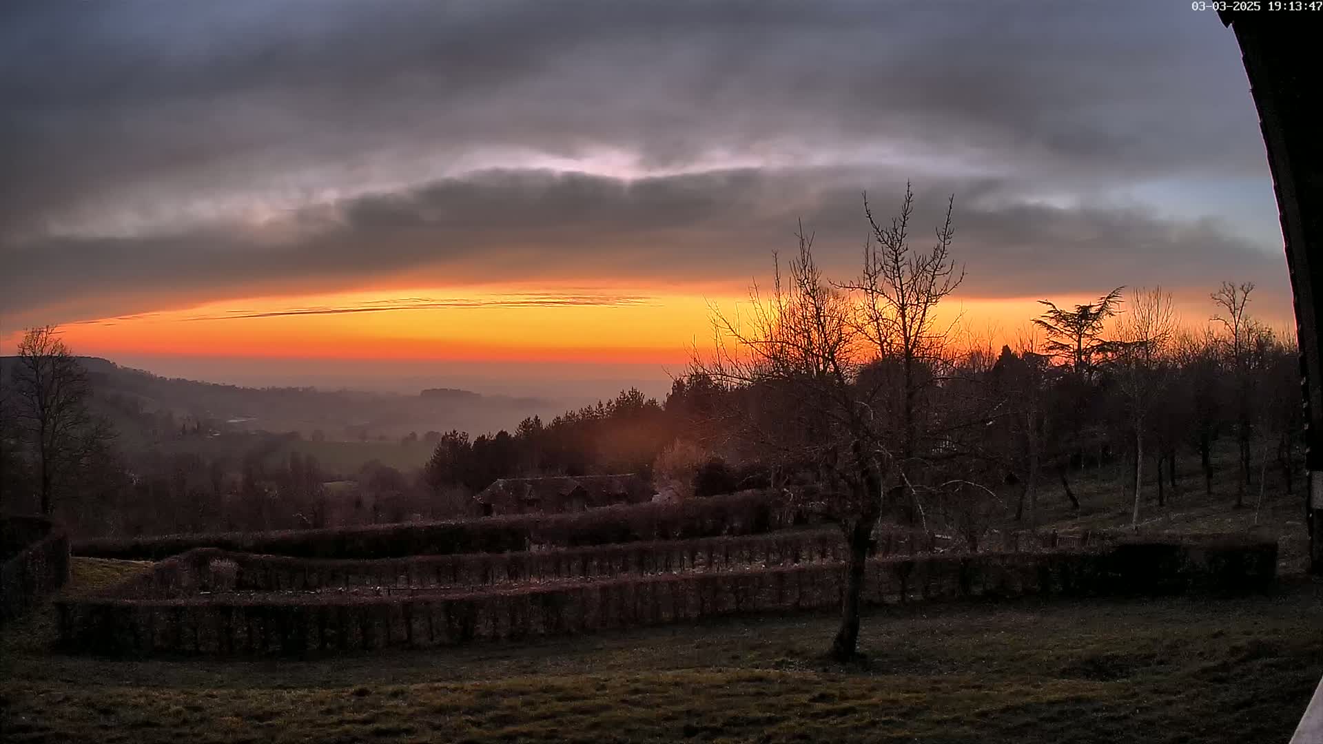 A vibrant orange sunset over a hazy valley, viewed from a hillside with leafless trees and neatly trimmed hedges in the foreground under a partly cloudy sky.