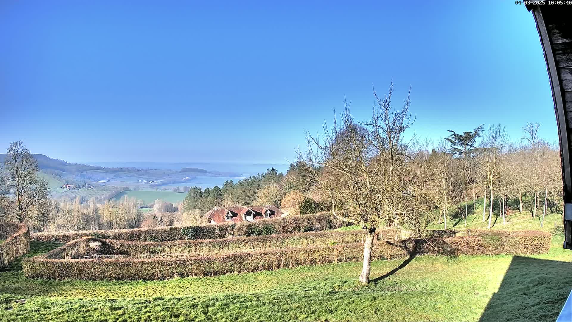 A sunny, clear day overlooks a landscape of rolling hills, trees, a building, and meticulously trimmed hedges, with a body of water visible in the distance.