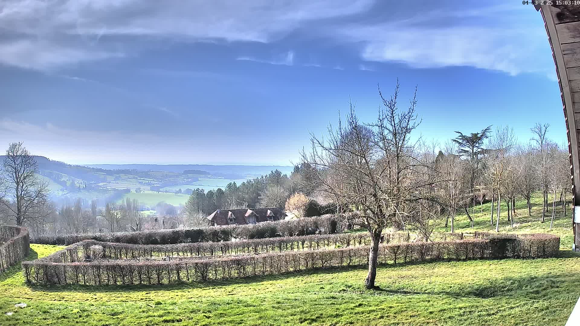 A partly cloudy blue sky overlooks a rural landscape with a building nestled behind neatly trimmed hedges, all situated on a grassy hillside.