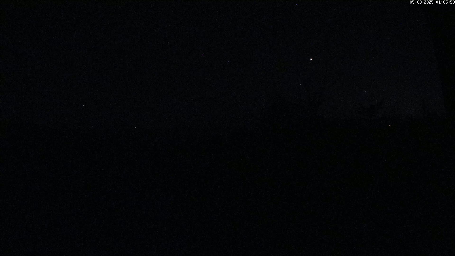 A nighttime, dark outdoor scene shows a few visible stars in a clear sky above a dark, silhouetted landscape.