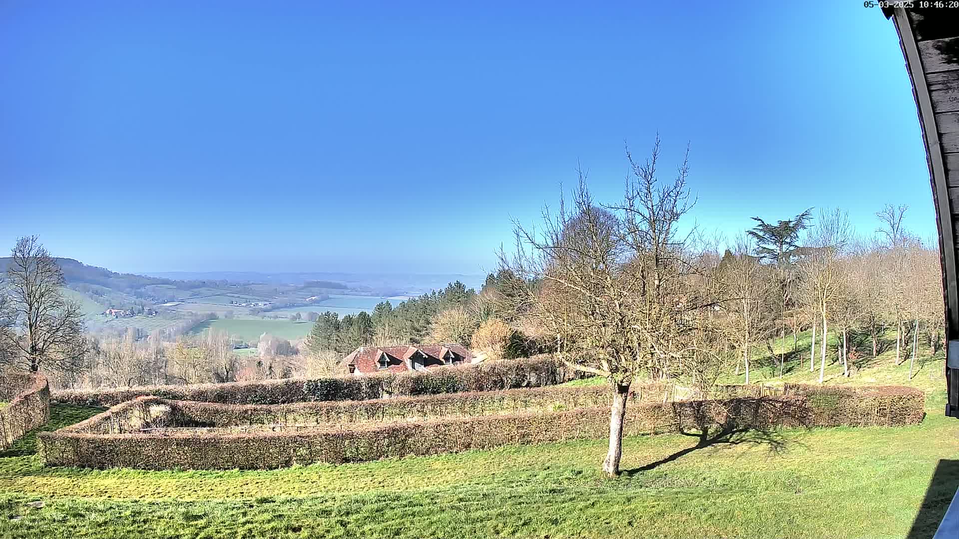 A sunny day reveals a picturesque valley view, showcasing a distant body of water, rolling hills, and a landscape dotted with trees and a building partially obscured by hedges.