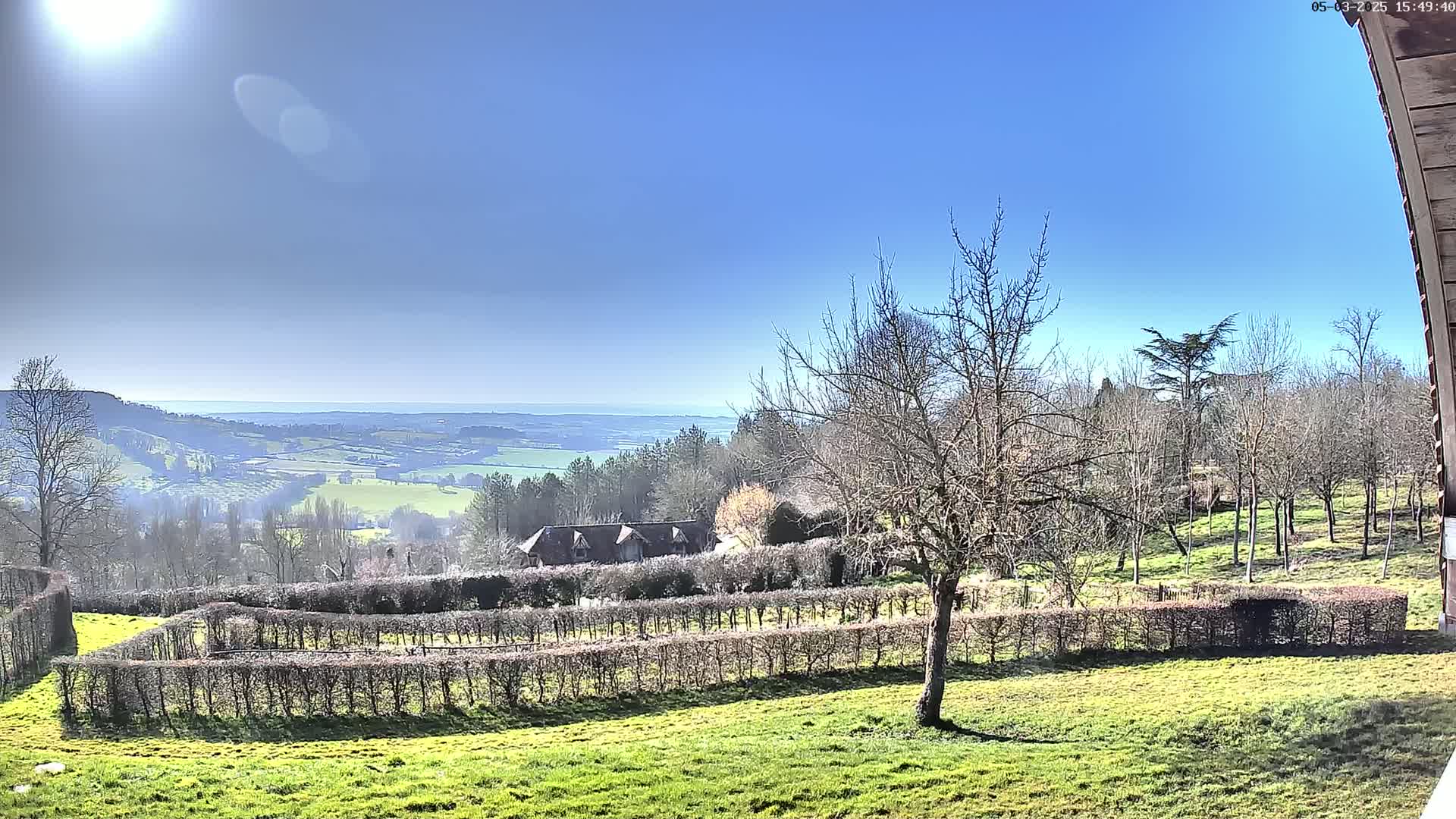 A sunny day reveals a panoramic view of a sprawling green valley, bordered by leafless trees and hedgerows, with a distant body of water visible under a clear blue sky.