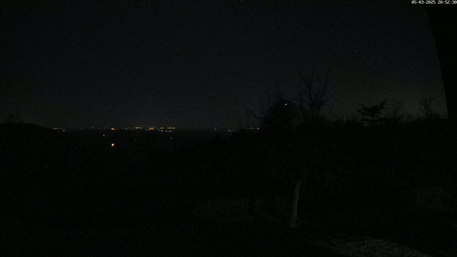 A dark nighttime scene shows a distant line of city lights across a valley, silhouetted against a mostly clear, star-speckled sky.