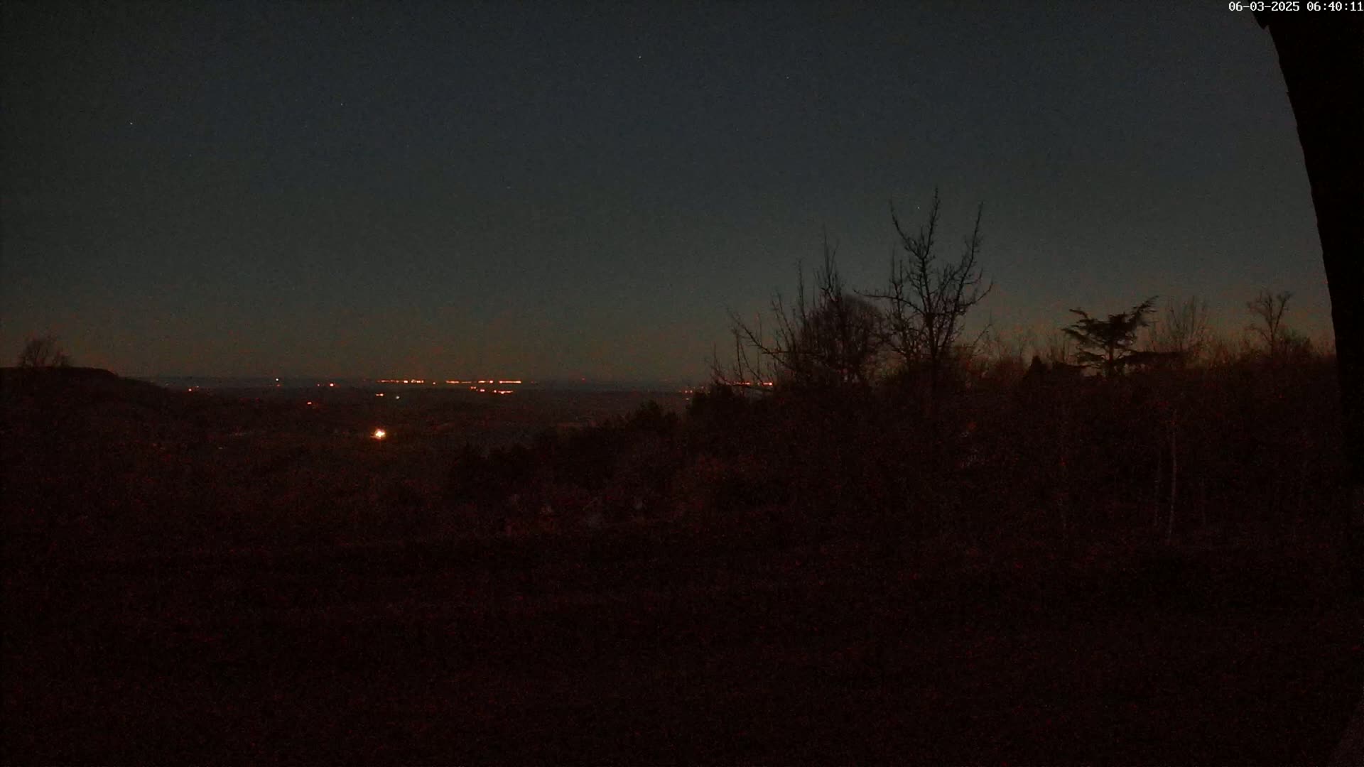 A dark, nighttime view of a hilly landscape with distant city lights under a mostly clear sky.