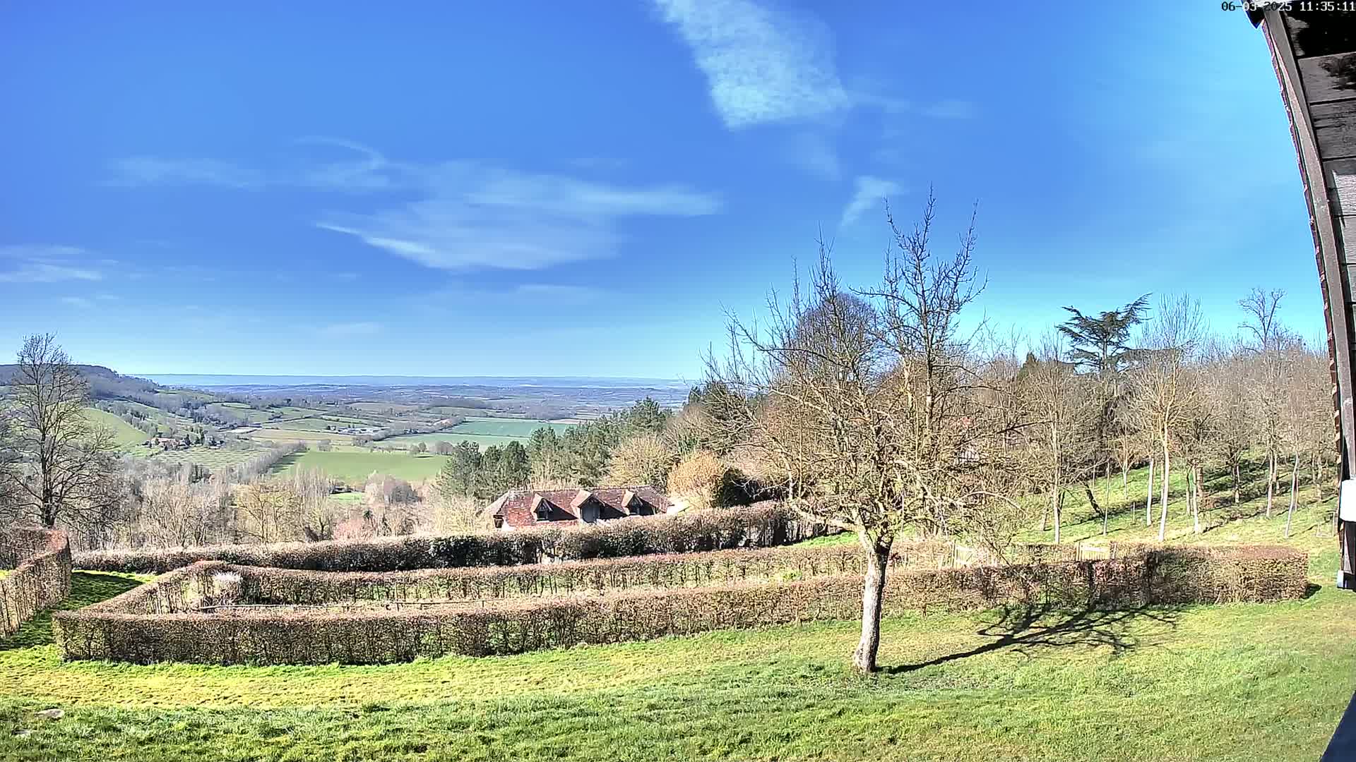 A sunny day reveals a sweeping vista of farmland and rolling hills, bordered by a neatly trimmed hedge surrounding a grassy yard and a building in the mid-ground.