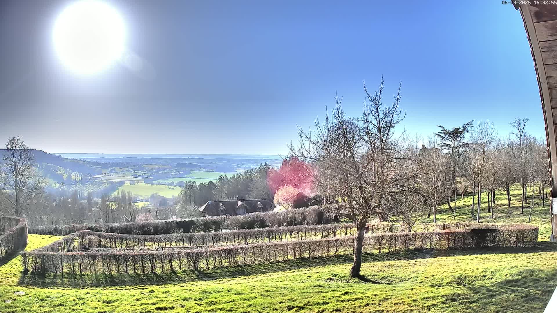 A sunny day reveals a vast, verdant valley viewed from a hillside, featuring a house nestled amongst meticulously trimmed hedges and leafless trees.