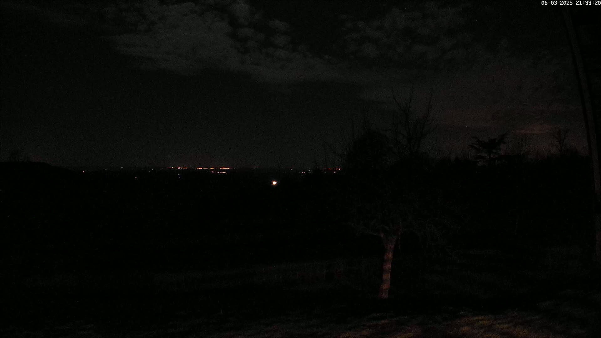 A nighttime view of a dark landscape with a few distant lights visible on the horizon under a cloudy sky.