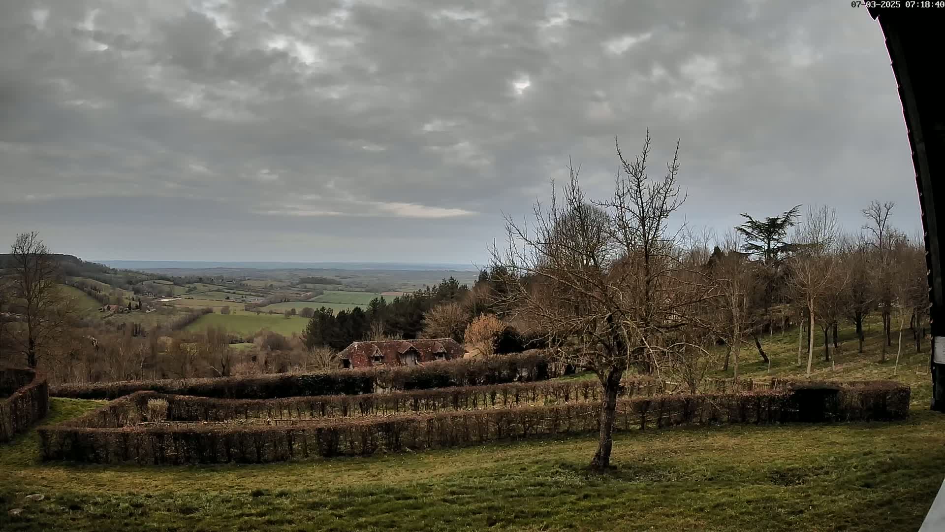 A sprawling, overcast landscape features a distant valley, a building nestled among bare trees and hedgerows in the midground, and a field in the foreground.