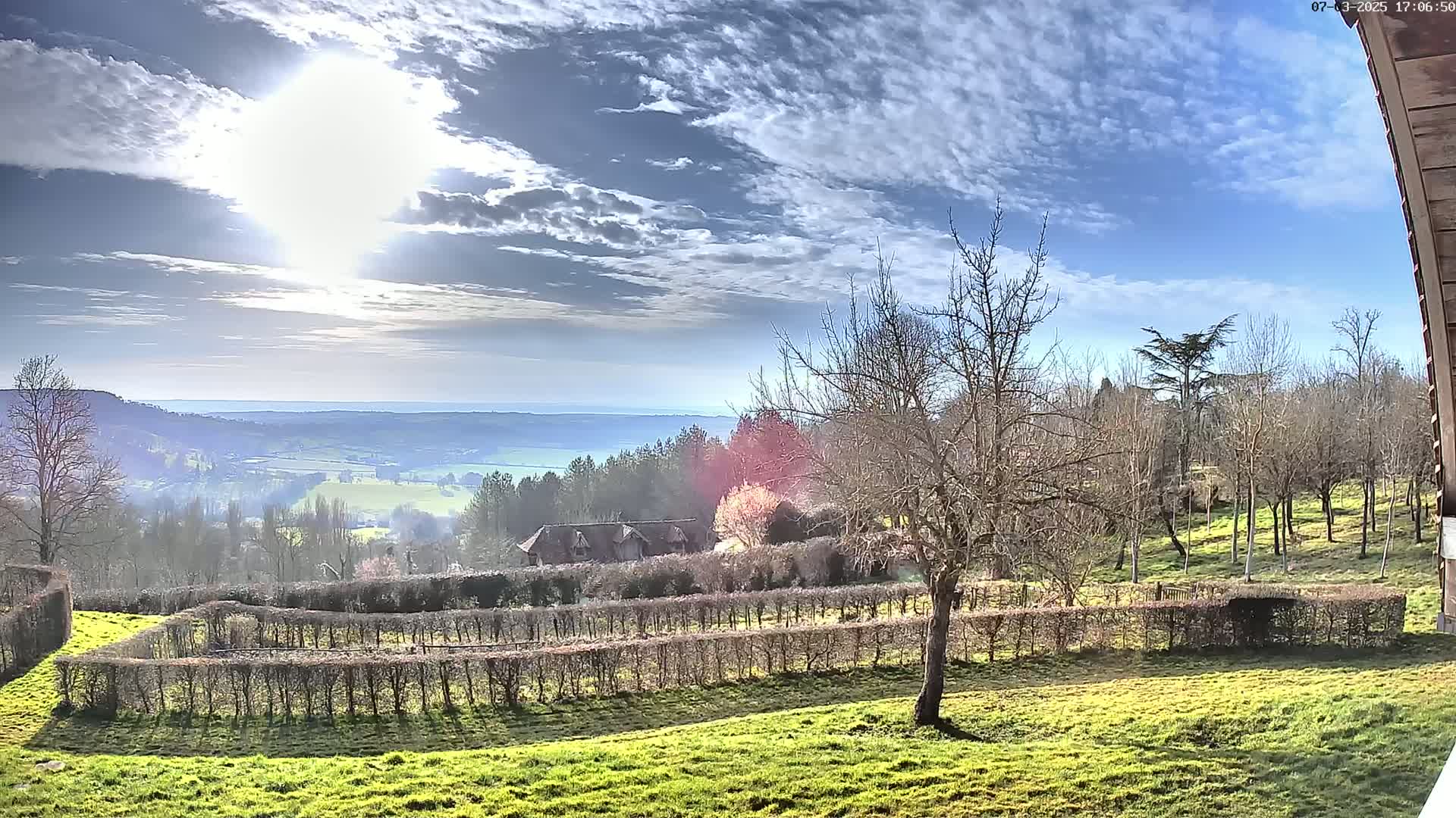 A sunny day reveals a sprawling green landscape with neatly trimmed hedges, a cluster of buildings nestled amongst trees, and a distant valley extending to a hazy horizon under a partly cloudy sky.