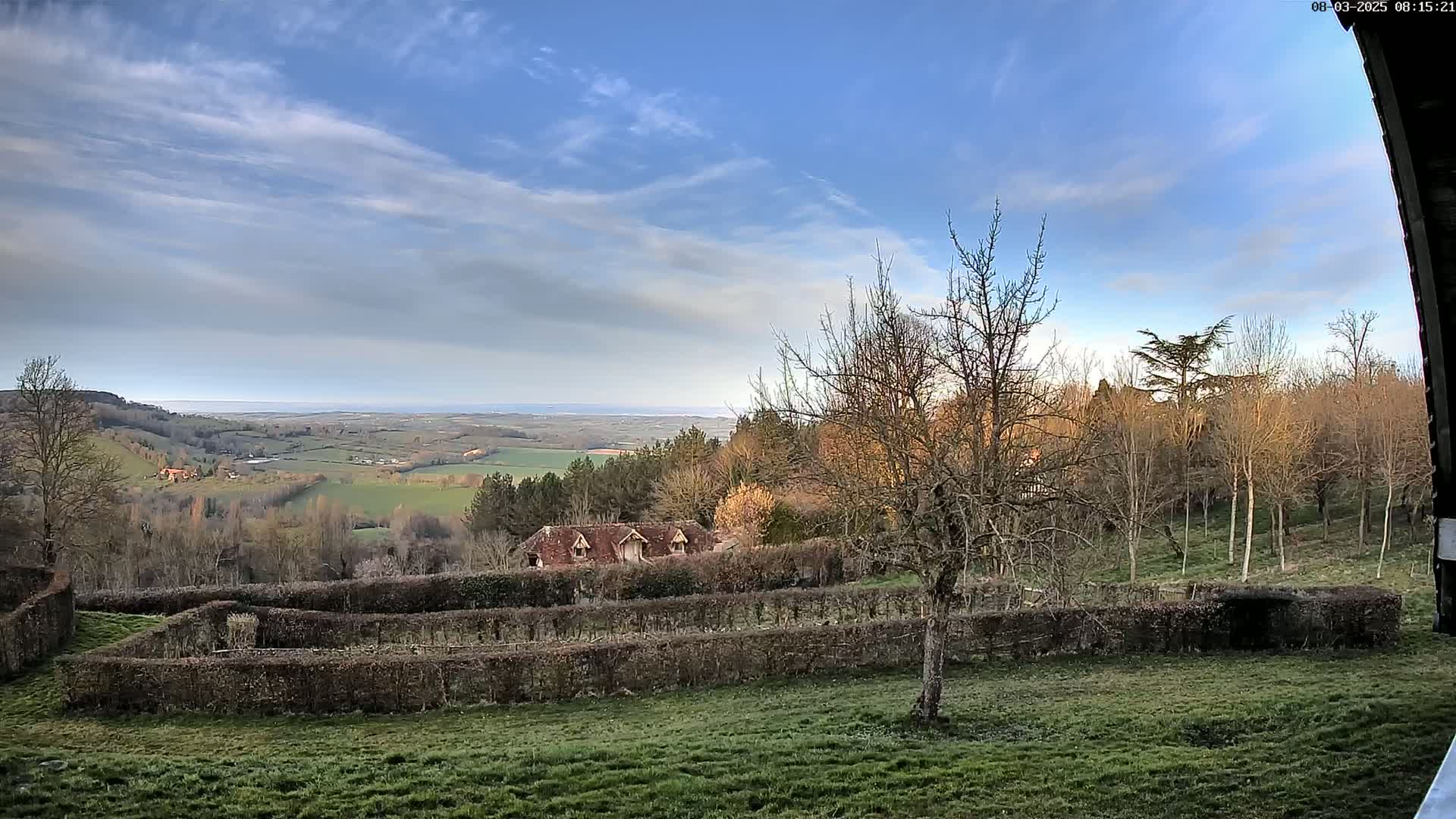 A partly cloudy blue sky overlooks a tranquil landscape of rolling green hills, a distant body of water, and a building nestled among bare trees and neatly trimmed hedges.