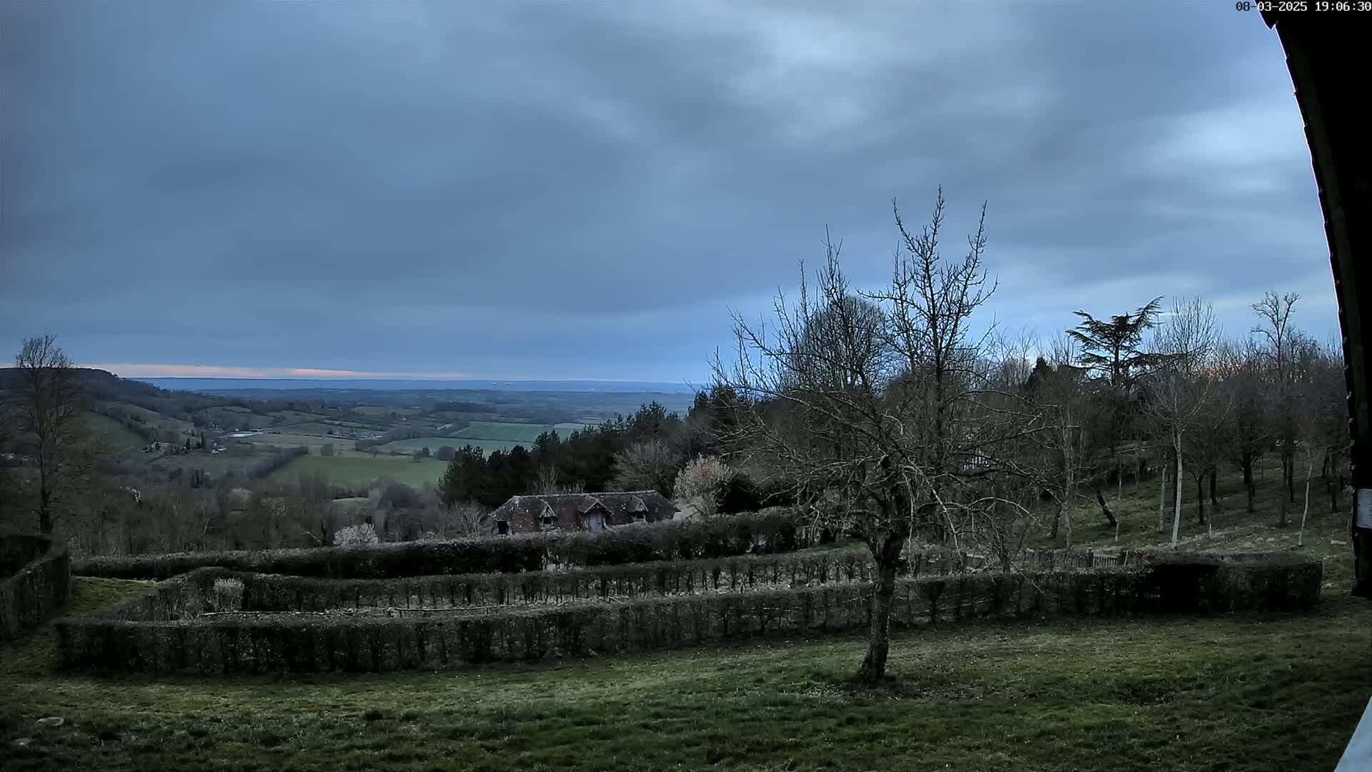 A sprawling, verdant valley stretches out under a cloudy, overcast sky, featuring a small building nestled among bare trees and neatly trimmed hedges in the foreground.