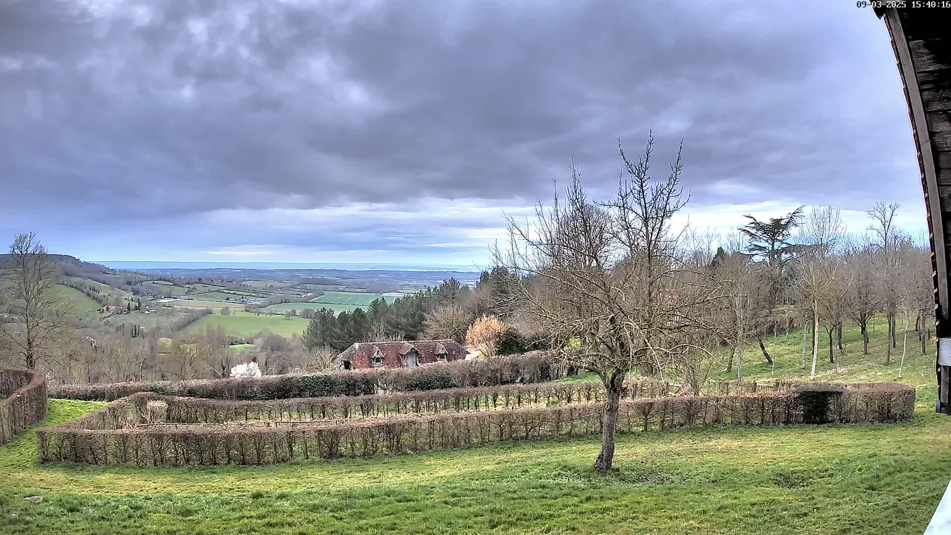 A sprawling, verdant valley under a cloudy sky stretches to a distant horizon, partially visible from behind a hedgerow surrounding a building and bare deciduous trees.