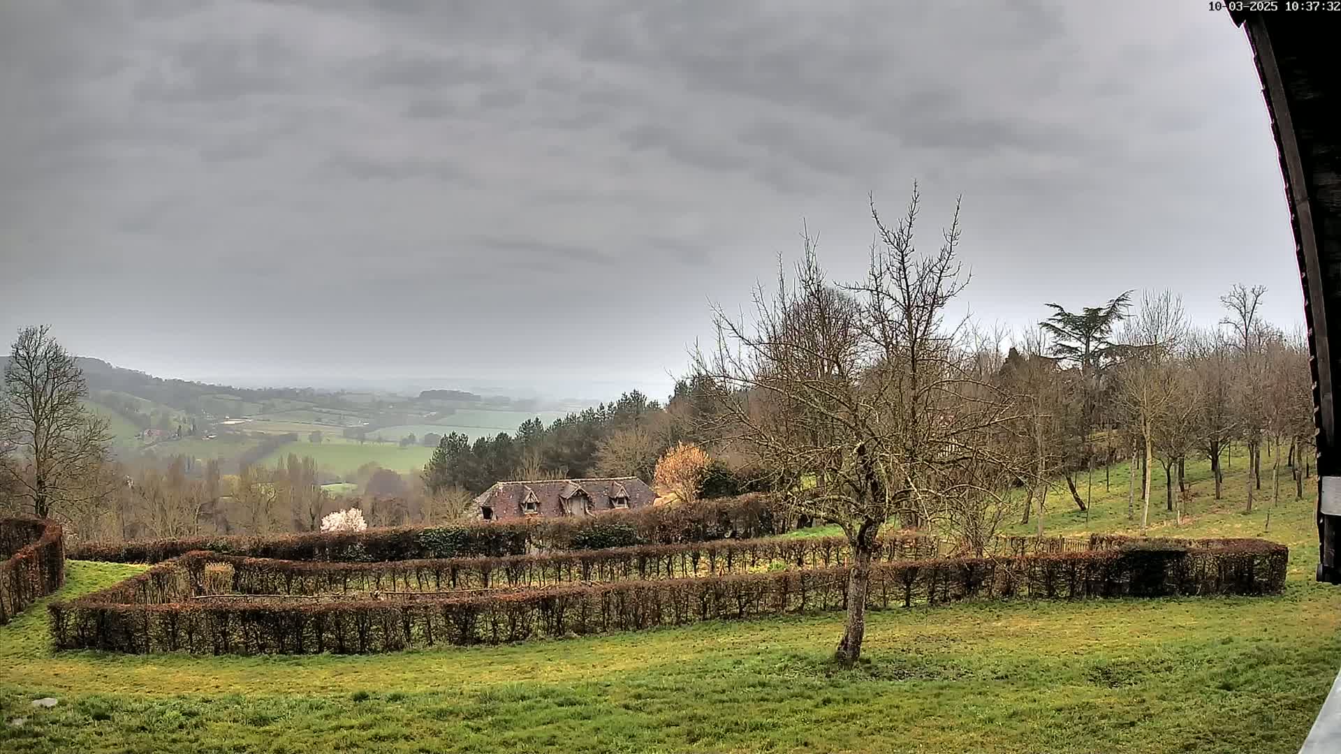 A sprawling, overcast landscape features a farmhouse nestled behind a meticulously manicured hedge, overlooking a verdant valley under a gray sky.