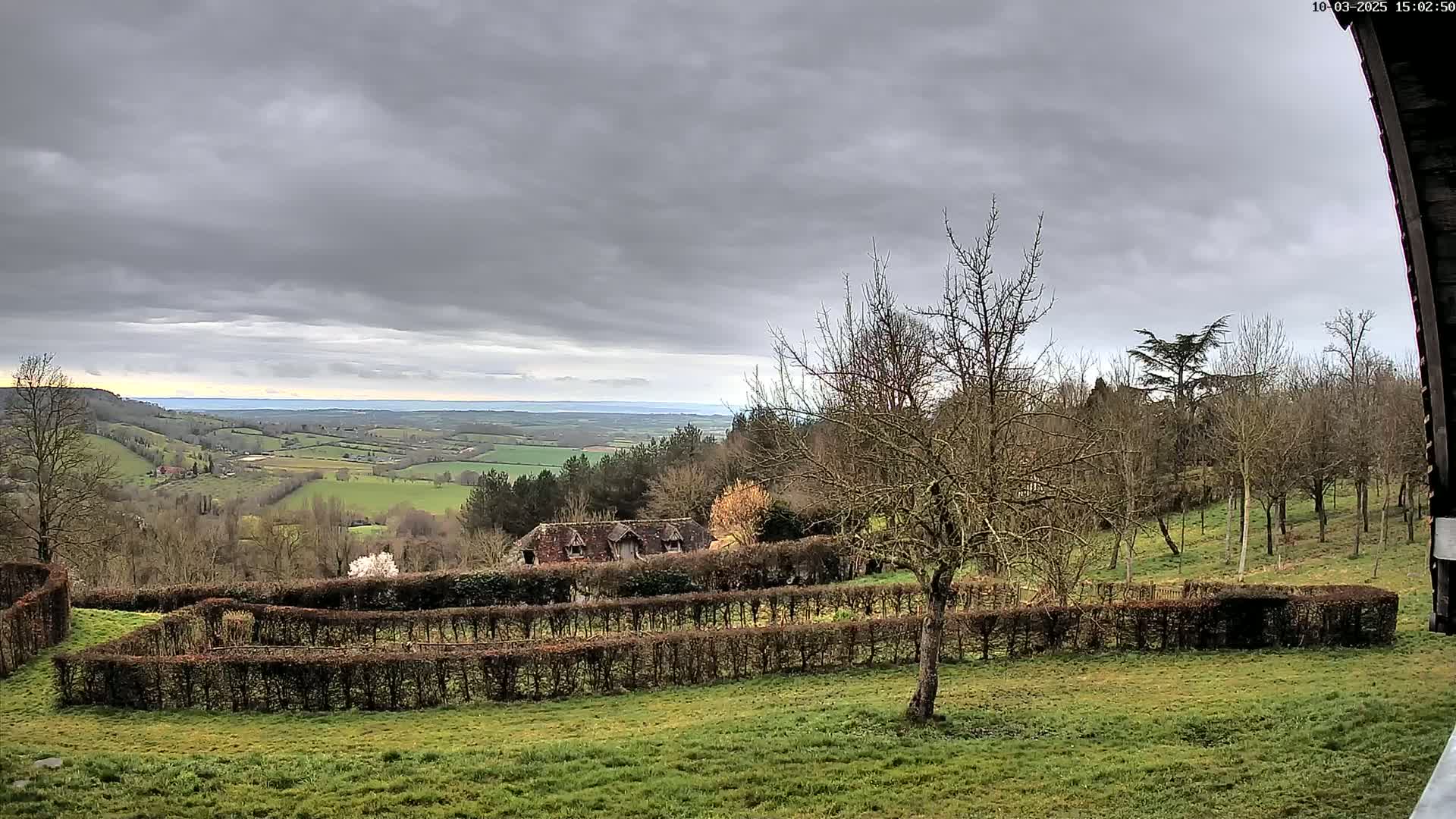 A sprawling green valley under a cloudy sky is visible from a vantage point overlooking a building partially obscured by hedges and bare trees.