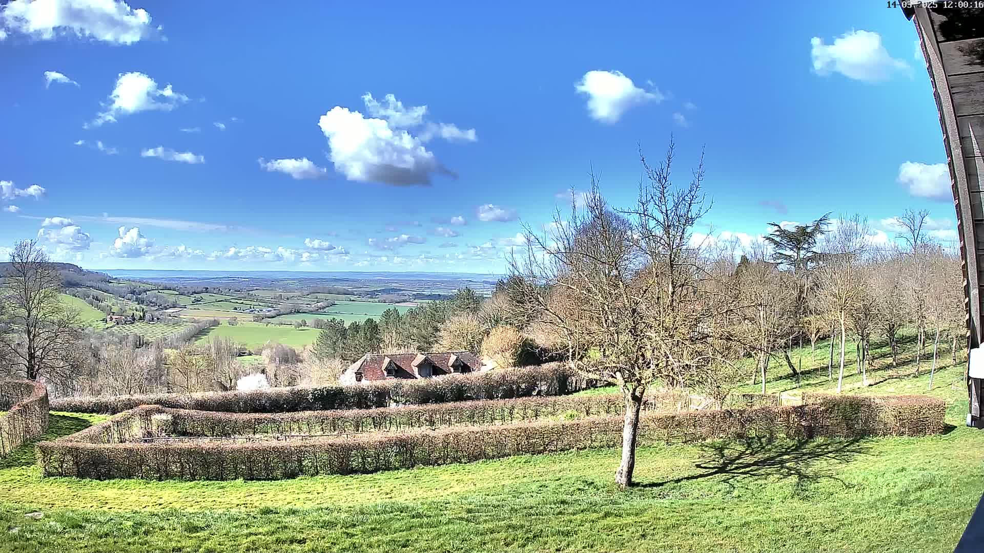 A sprawling green landscape under a mostly sunny sky with fluffy white clouds features a building nestled among trees and hedges, overlooking a distant valley.
