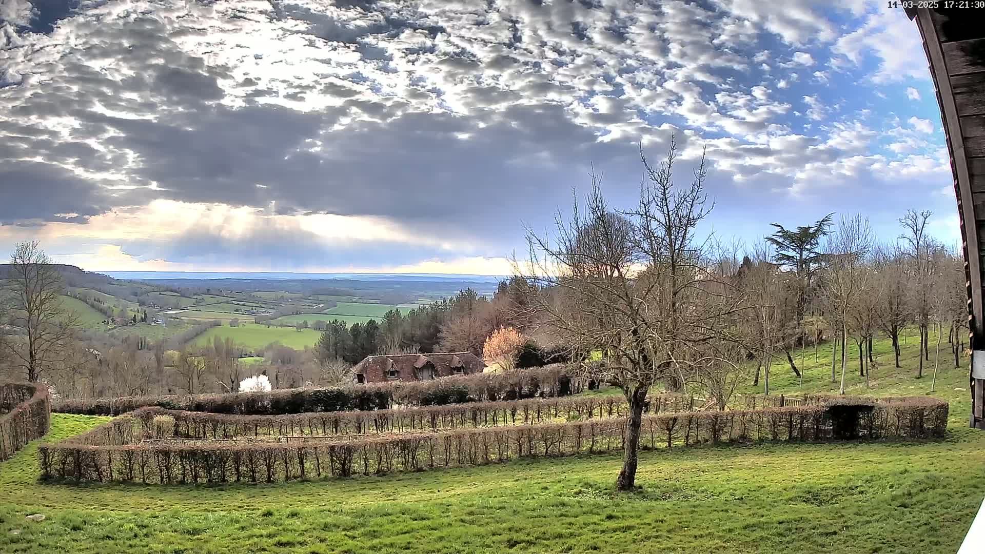 A partly cloudy sky brightens a pastoral valley scene, showing a building nestled behind neatly trimmed hedges and a stand of leafless trees.