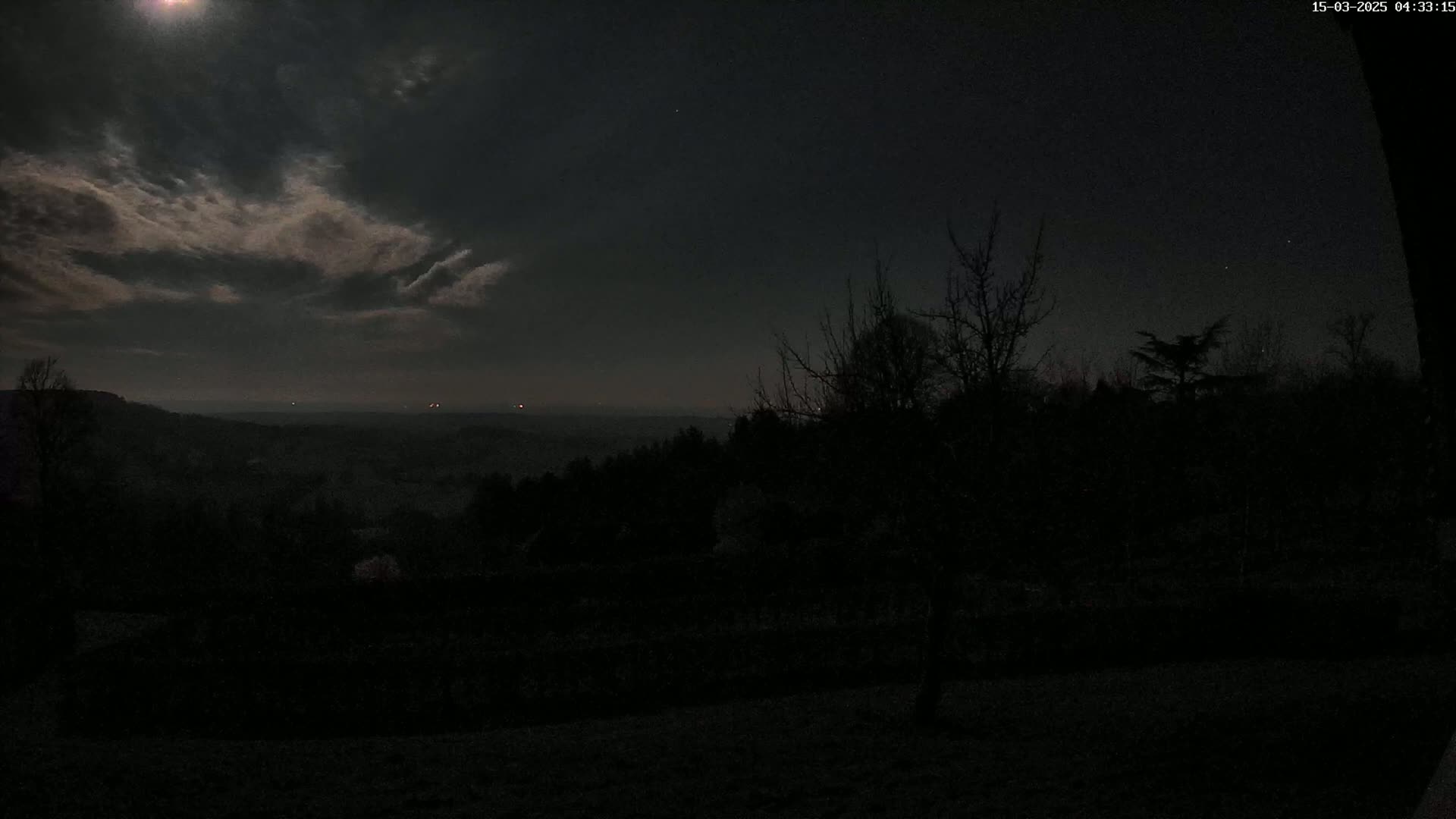 A nighttime view of a dark, wooded landscape under a mostly cloudy moonlit sky.