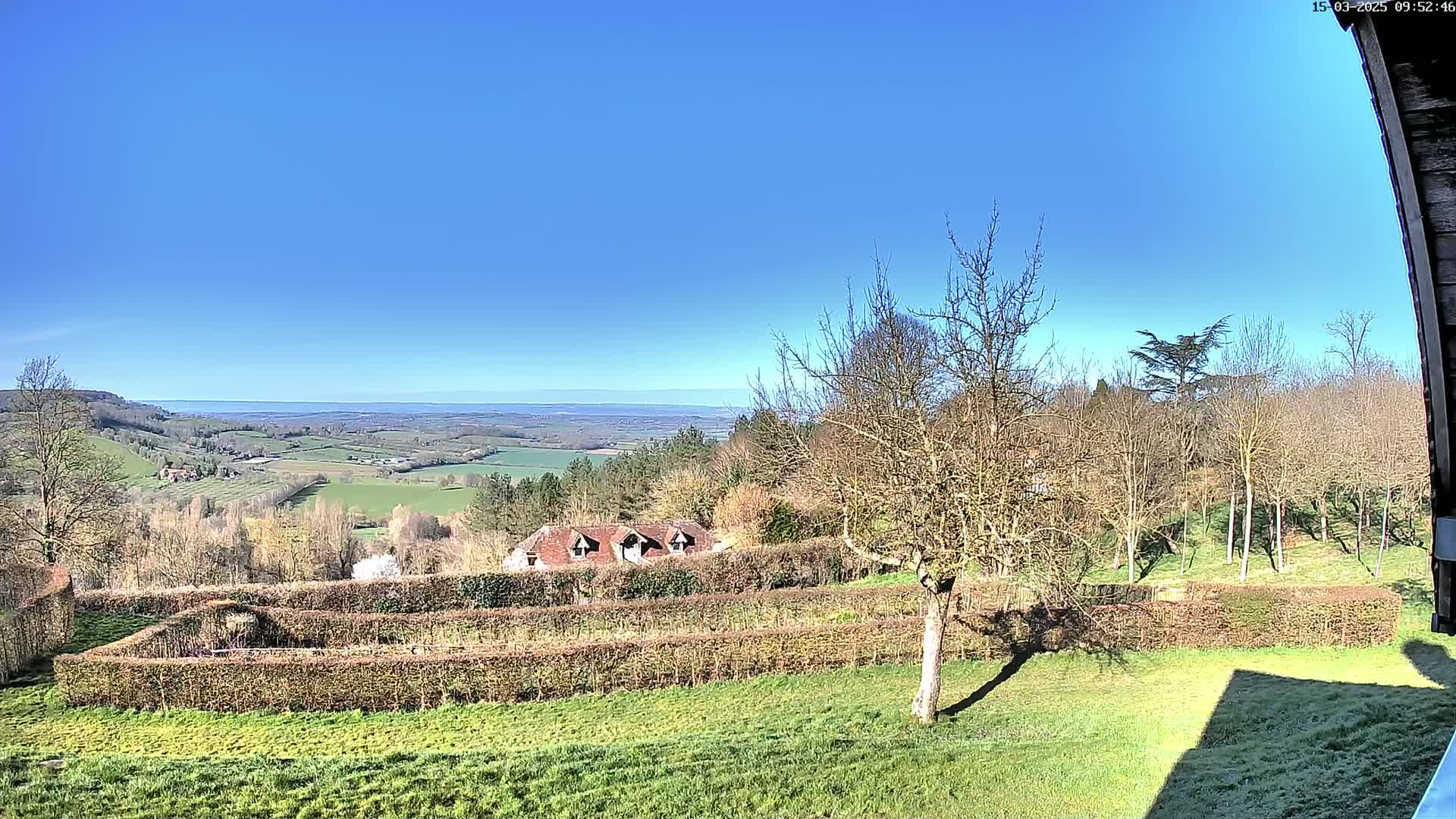 A sunny, clear day reveals a panoramic view of a rolling green landscape, featuring a house nestled amongst trees and hedges, with a distant horizon visible.