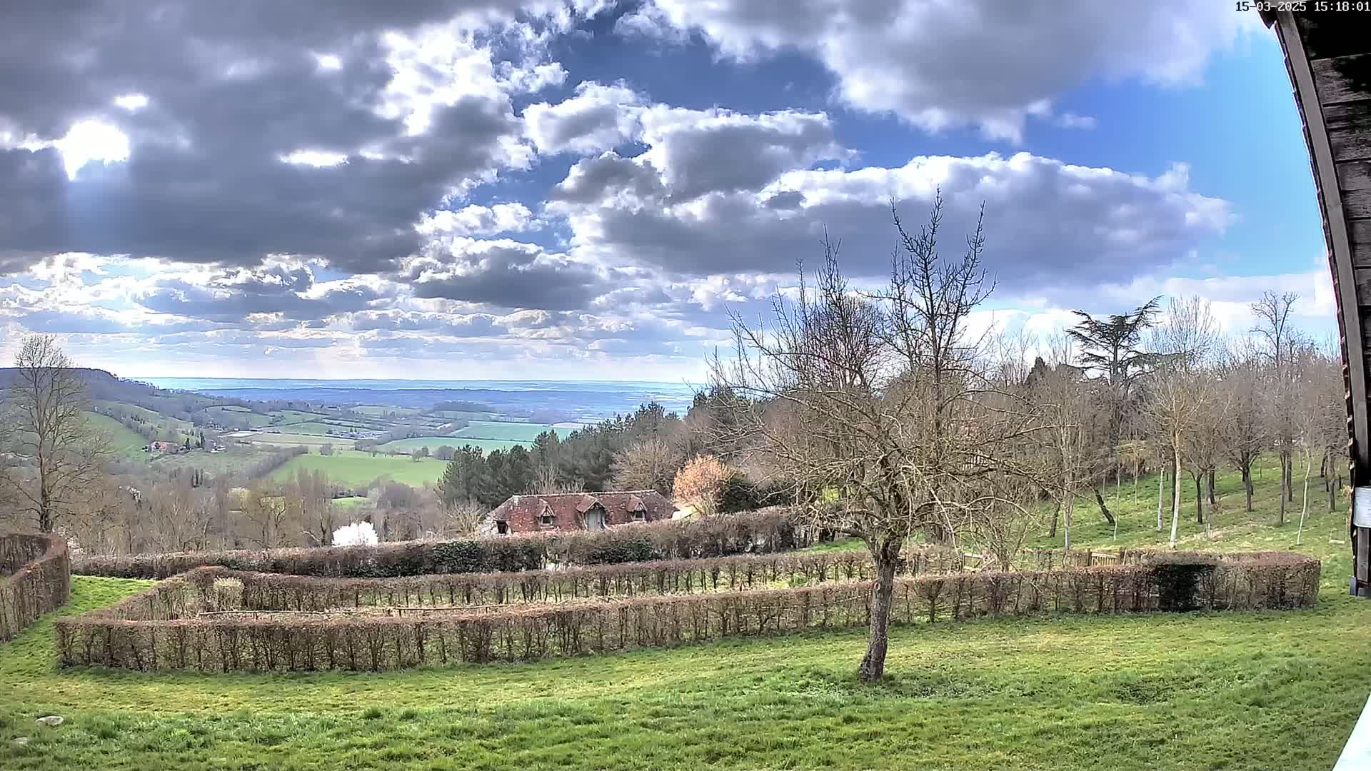 A partly cloudy day reveals a rolling green landscape with a building nestled amongst trees and hedges, overlooking a distant body of water.