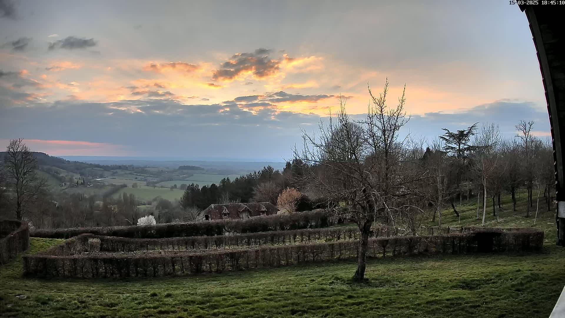 A picturesque sunset over a sprawling green valley, viewed from a hillside with a building and hedged gardens in the foreground, under a partly cloudy sky.