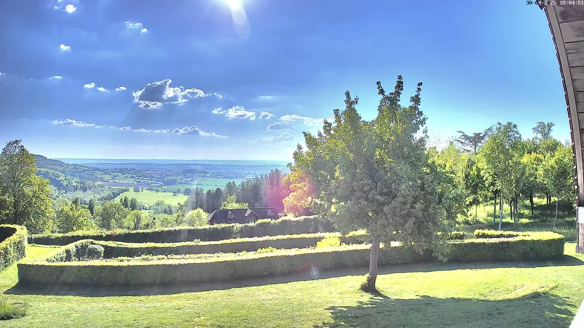 A sunny day reveals a sprawling green landscape, featuring neatly trimmed hedges, a tree in the foreground, and a distant view of rolling hills and a valley under a partly cloudy sky.