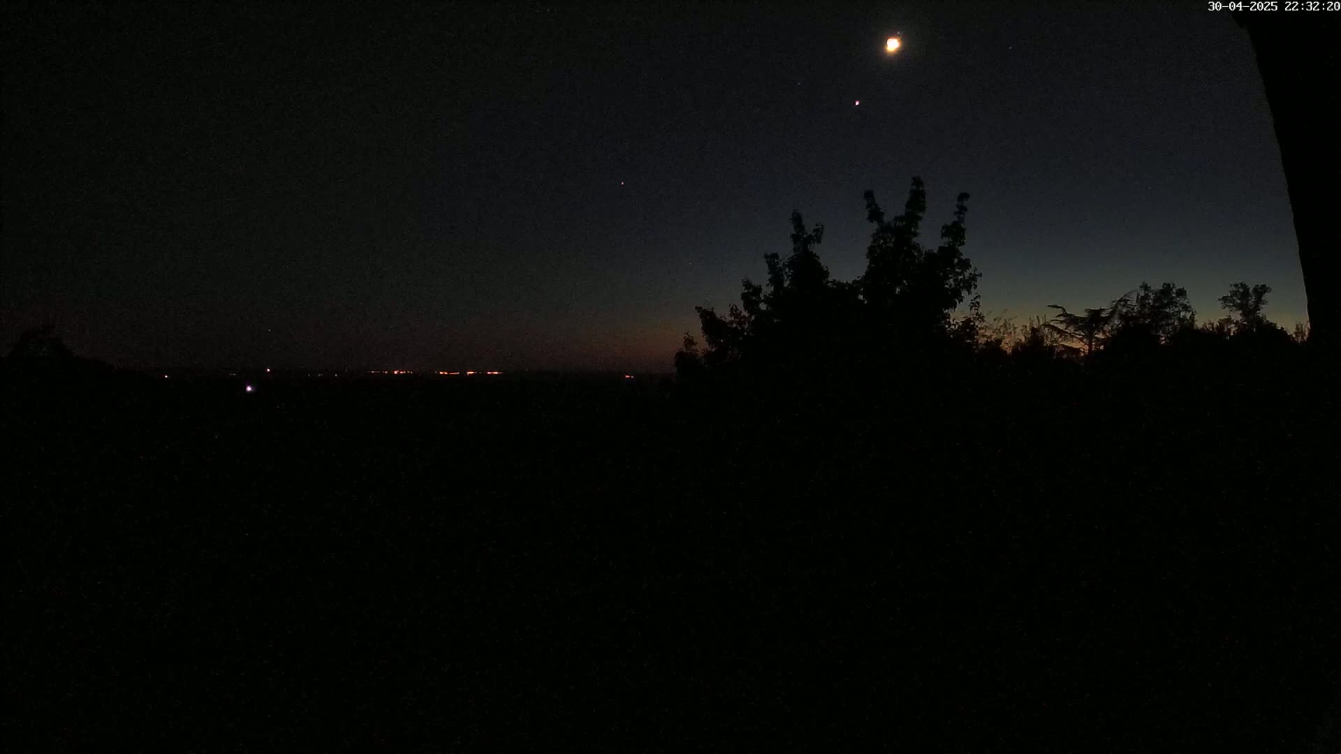 A nighttime scene shows a crescent moon and stars in the dark sky above a silhouetted landscape of trees and distant lights.